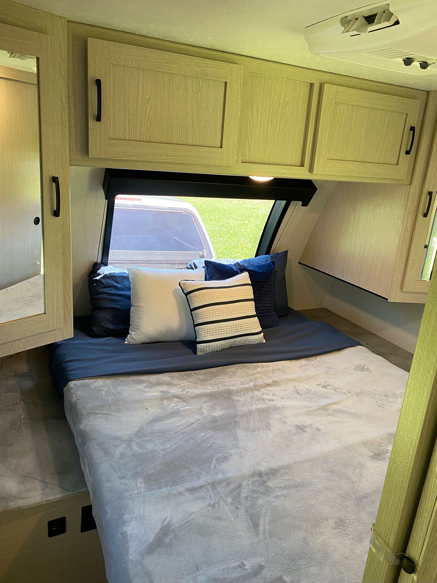 RV rental interior: A cozy bedroom with blue and grey bedding, various pillows, wooden cabinets, and a window view outside.. Forest River two bedroom Apex ultralite 2022