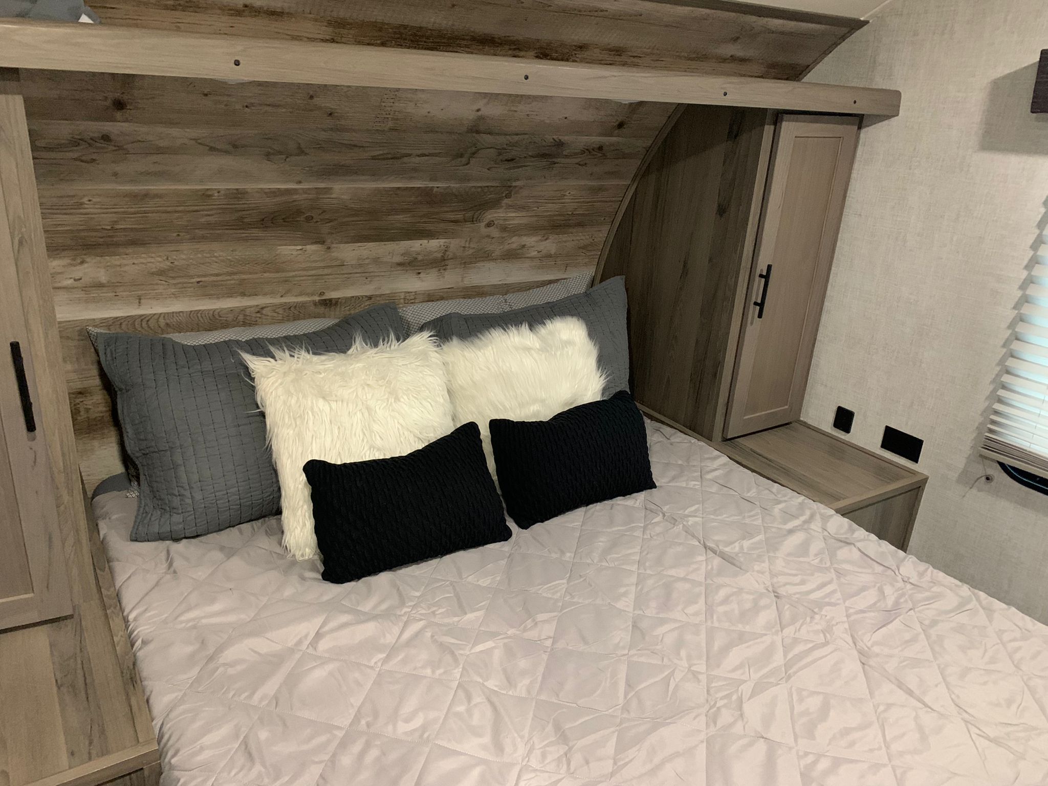 This RV rental bedroom features a comfy bed with grey quilt, fluffy white & black pillows, and rustic wood cabinetry.. Gulf Stream Ameri-Lite 2021