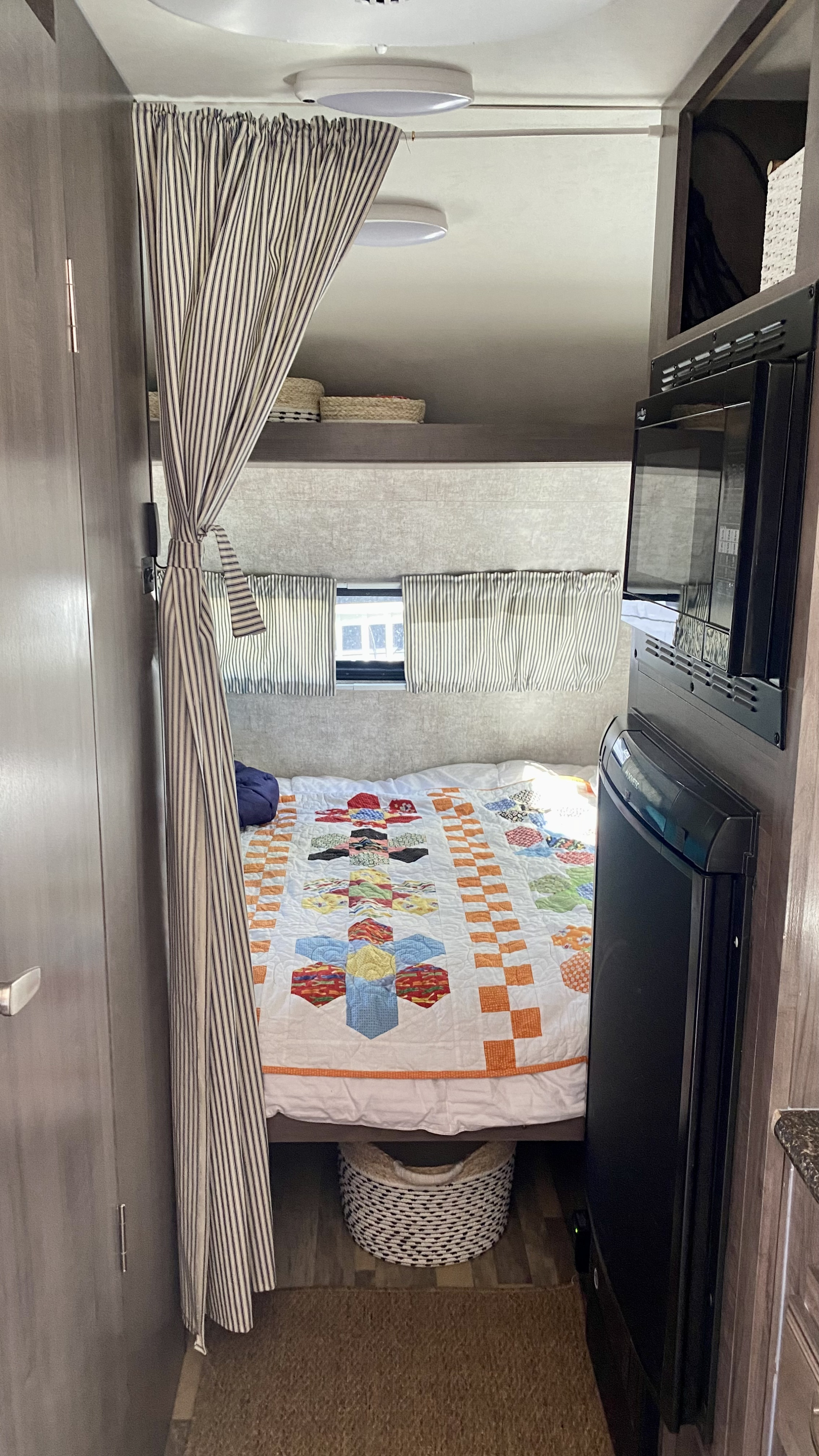 Cozy RV rental interior showing a bed with a colorful quilt, privacy curtain, and compact kitchen appliances.. Winnie Drop Winnie Drop Trailer 2018