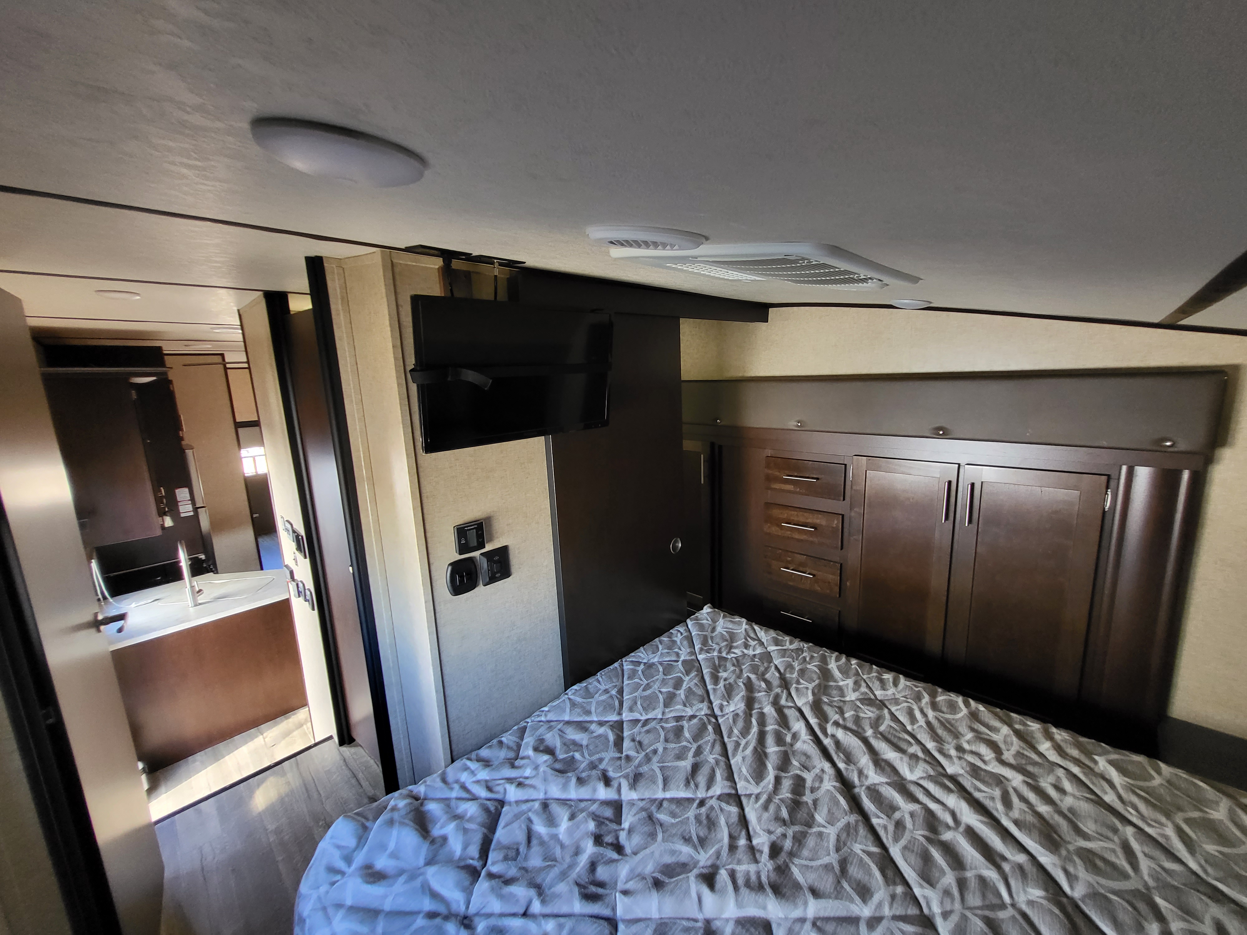 Inside a modern RV rental: a bedroom with a patterned bed, TV, and built-in storage. Kitchen visible beyond.. Forest River Cherokee Arctic Wolf 2018
