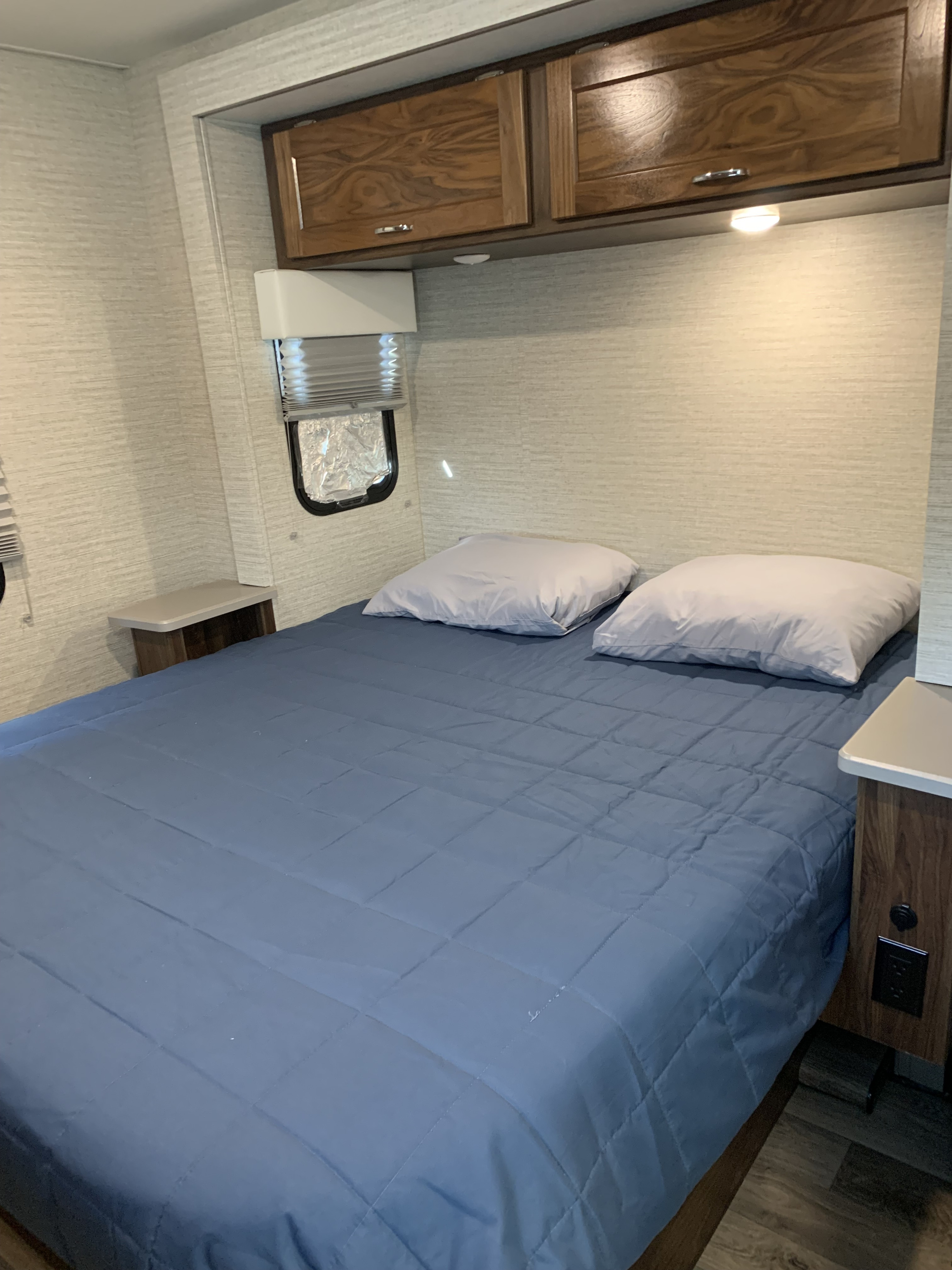 RV rental bedroom with a blue quilted bed, light pillows, overhead cabinets, and side tables for a comfortable stay.. Winnebago Outlook 2020