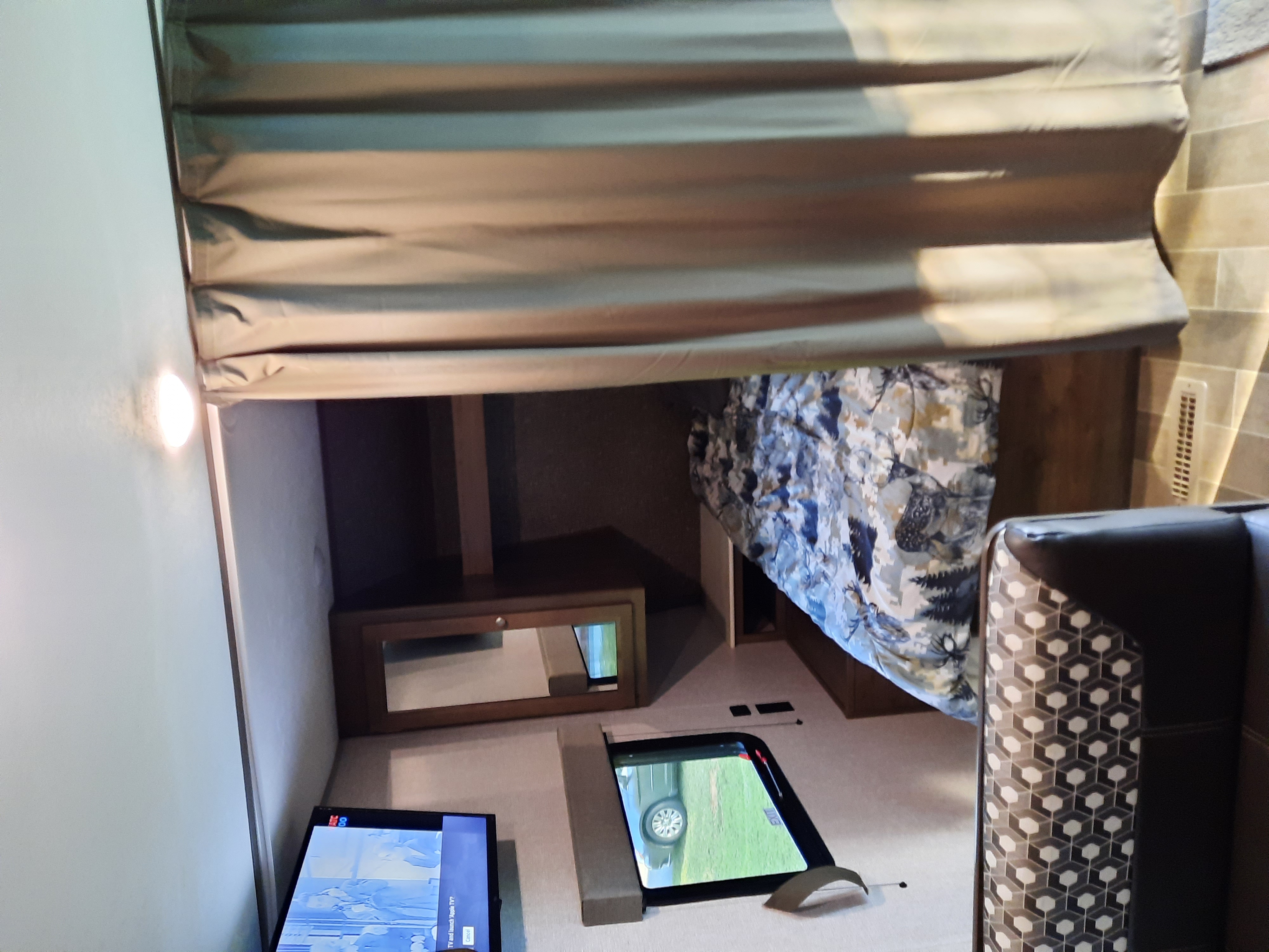 Inside an RV rental, a bunk area with a privacy curtain, patterned bedding, a small mirror, a TV, and a window view.. Dutchmen Coleman Lantern LT 2020