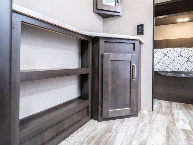 This RV rental showcases efficient storage with dark wood shelves, a cabinet, and a view of the compact bedroom.. Grand Design Transcend Xplor 2021