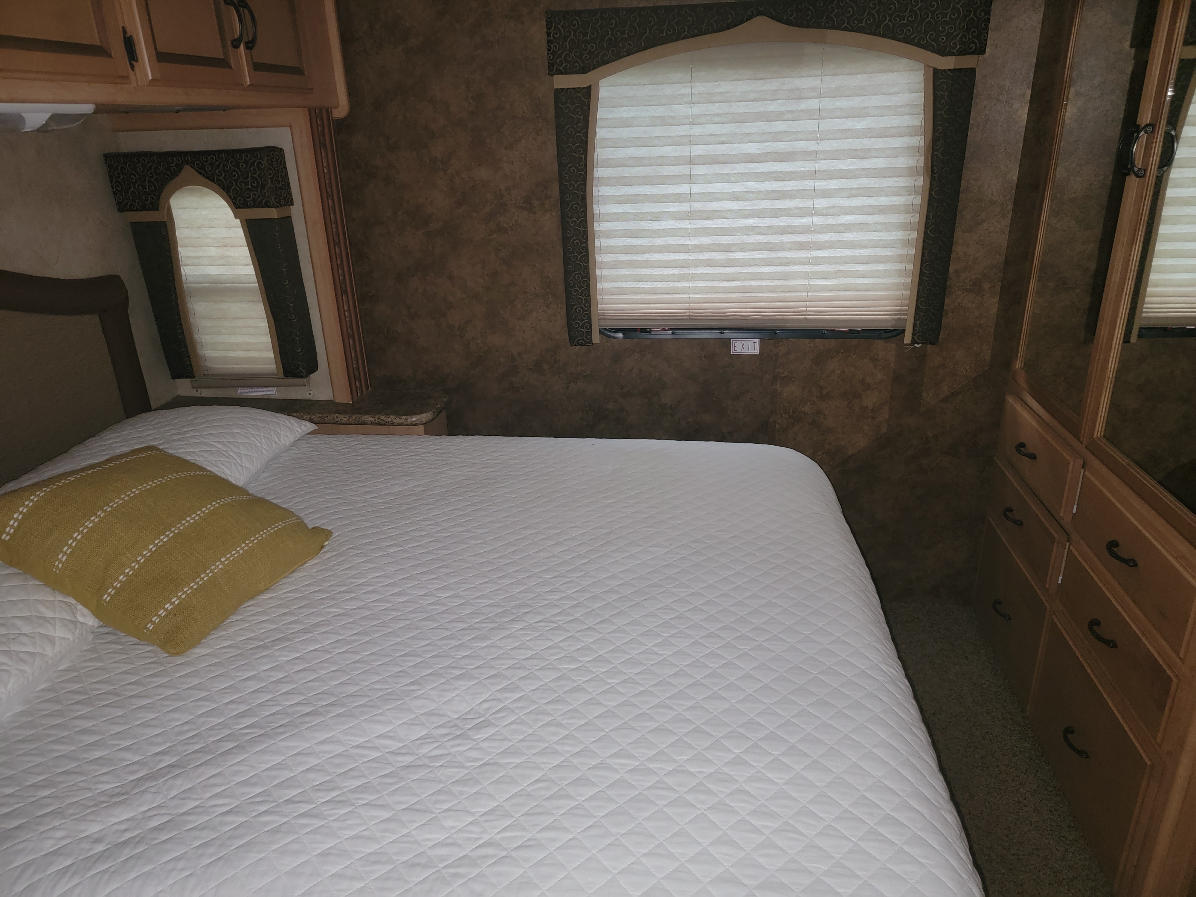 Comfortable RV rental bedroom with a white quilted bed, yellow pillow, wood cabinetry, and windows showing an 'EXIT' sign.. Thor Hurricane 2012