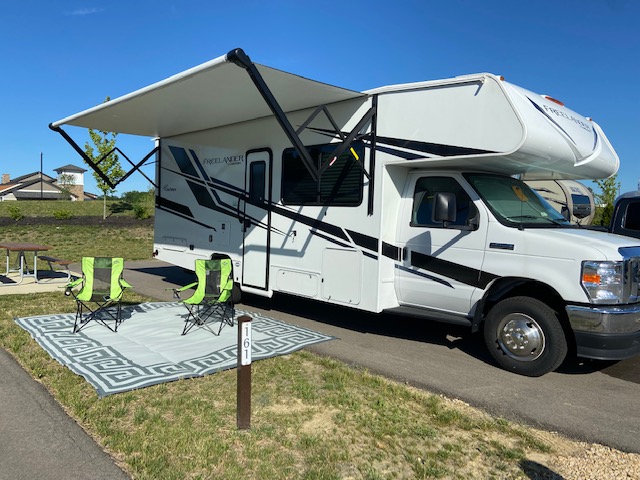 White Class C RV rental set up at a sunny campsite with extended awning, two green chairs, and an outdoor rug.. Forest River Coachmen Freelander Premier 2022