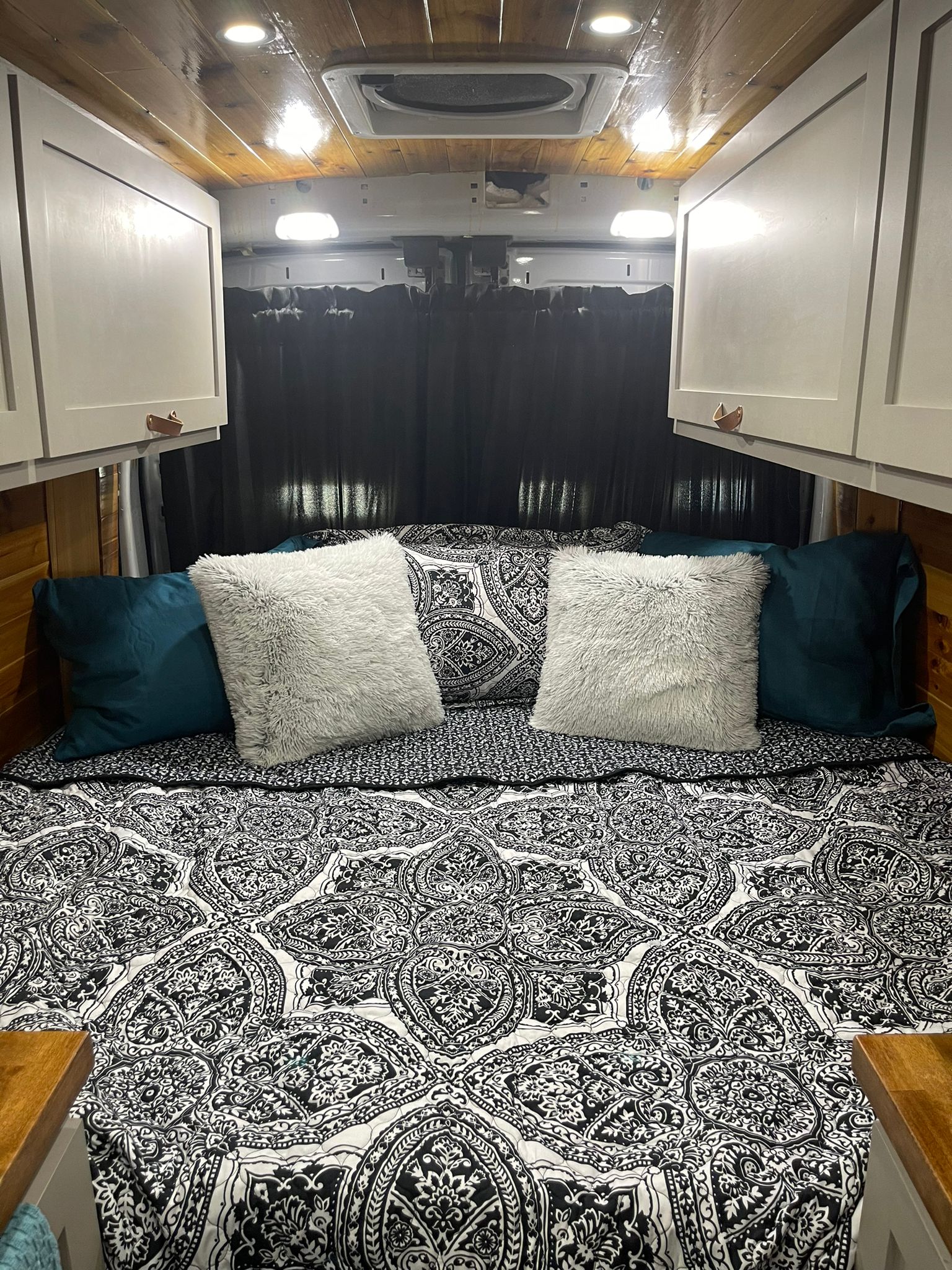 Inside this RV rental: a cozy bed with black/white patterned quilt, teal & fuzzy pillows, overhead cabinets.. Ford transit 2019