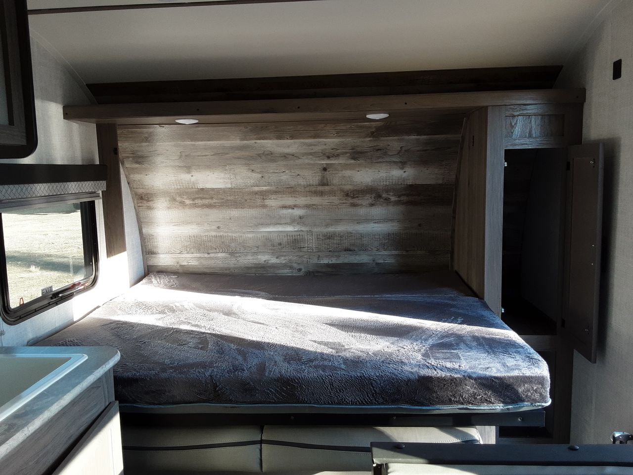 RV rental interior showing a compact bed with a cozy blanket, wooden accent wall, built-in storage, and a window.. Gulf Stream Kingsport 2022