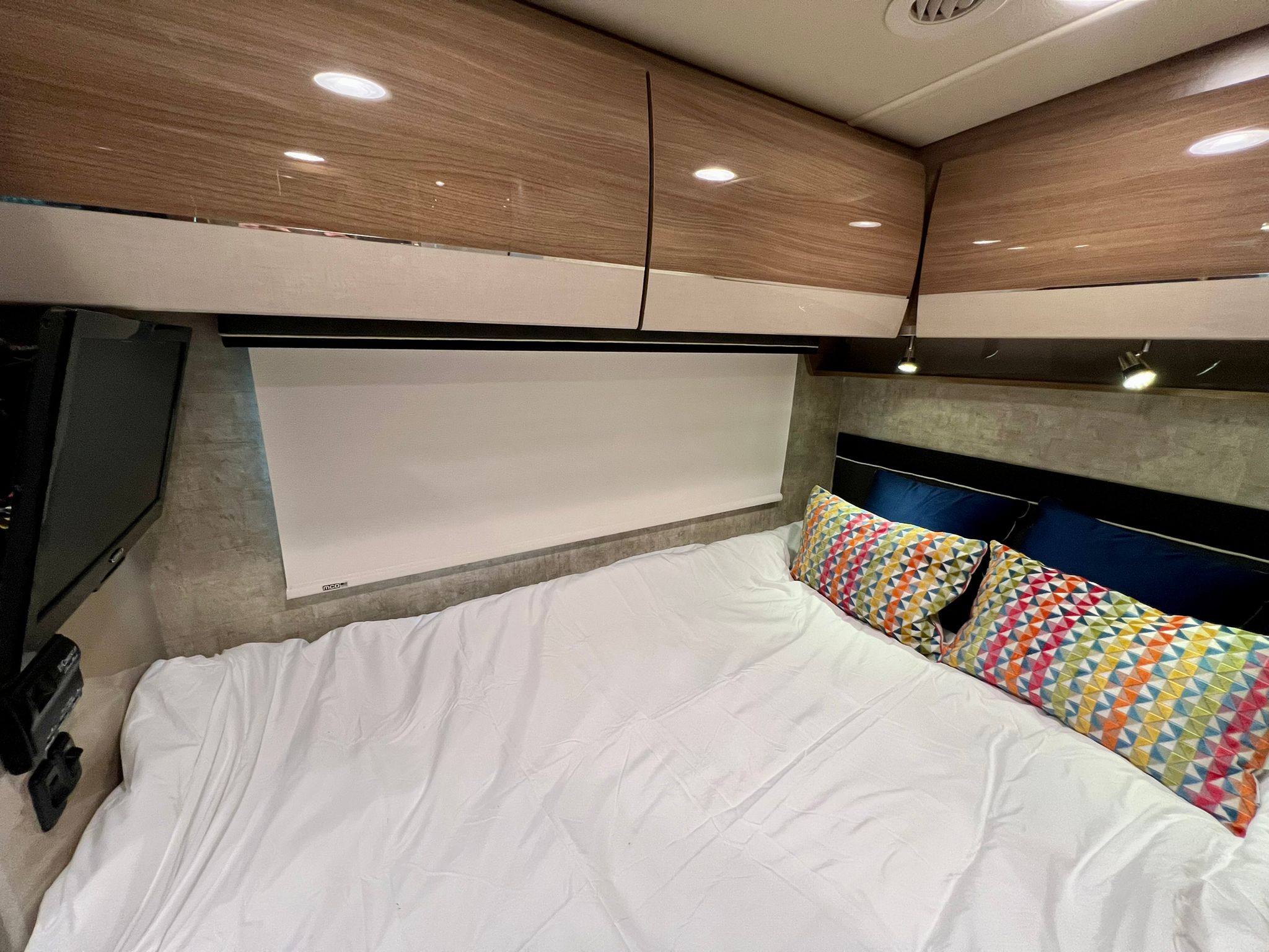 Modern RV rental bedroom with a comfy bed, colorful pillows, overhead cabinets, and TV.. Itasca Navion 2015