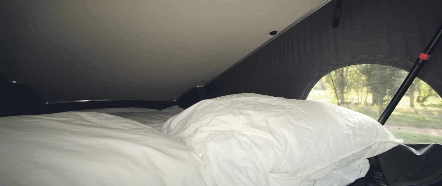 Cozy RV rental bed with white pillows and a view of trees. Perfect for camping adventures.. Ford Nugget L1 2022