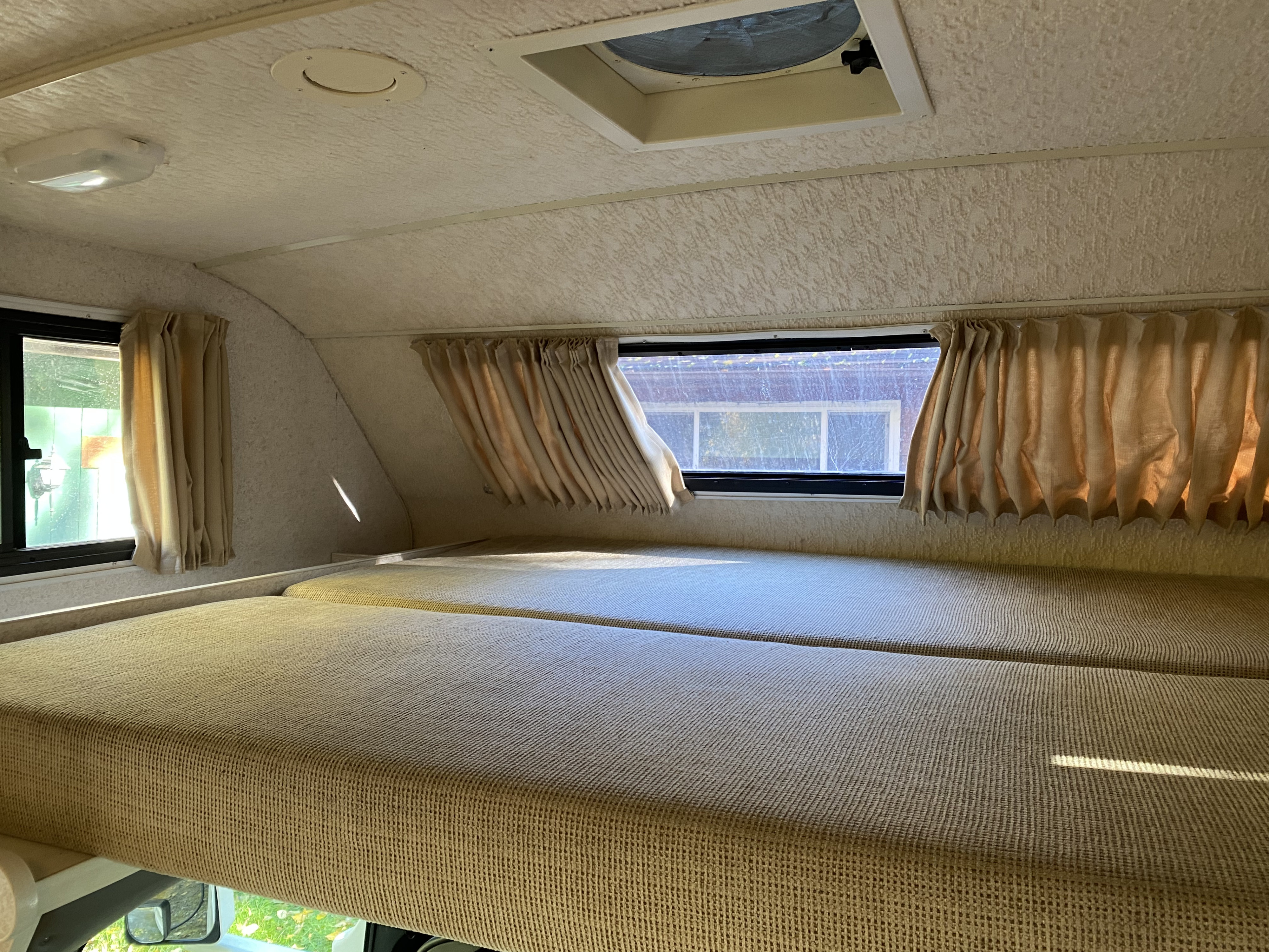 Inside an RV rental: a cozy cab-over bunk with two mattresses, side windows, and curtains. Ideal for a camper's restful night.. Winnebago Minnie 2004