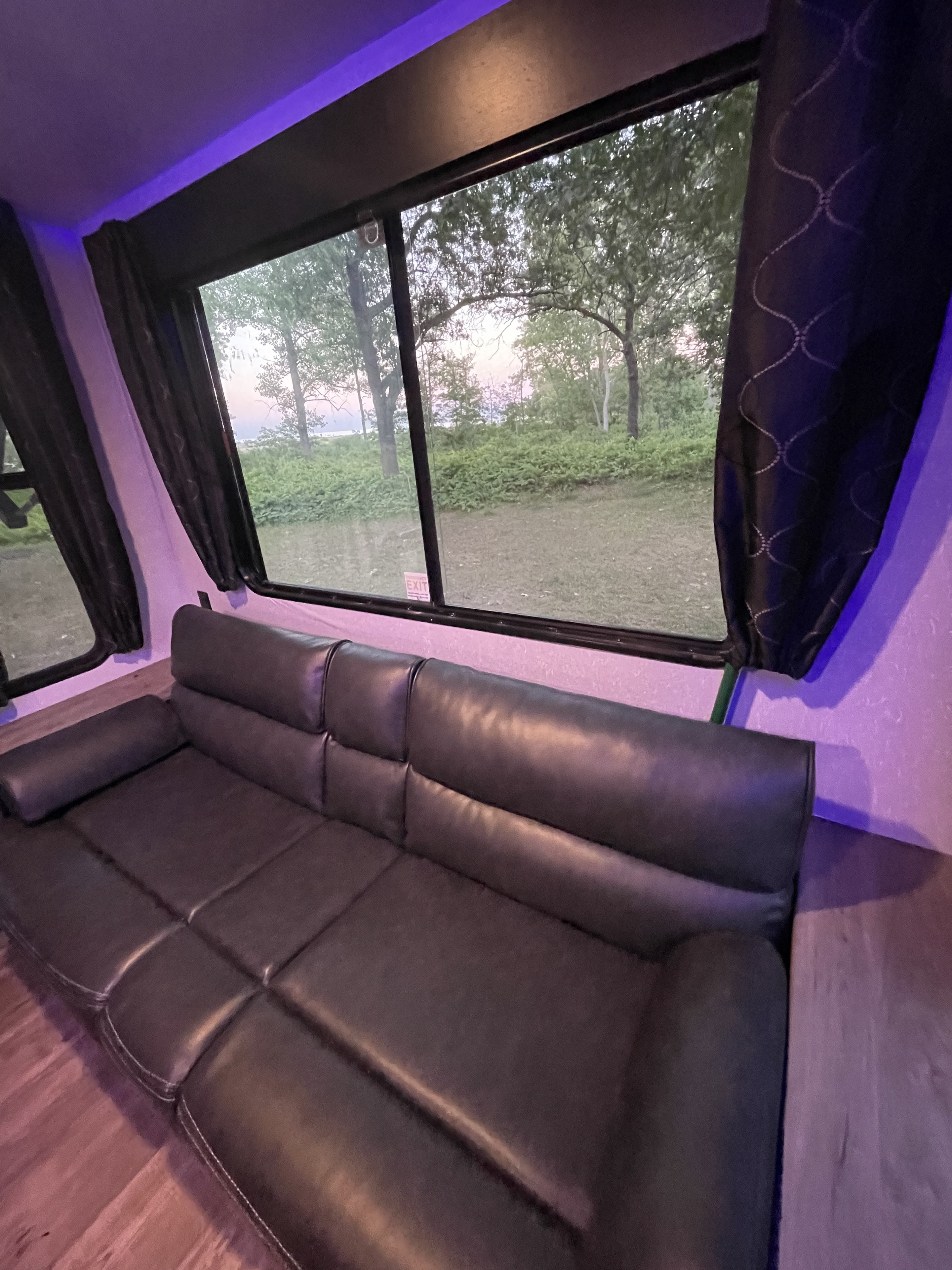 Inside a cozy RV rental: a plush black leather sofa illuminated by purple accent lights, offering a serene view of trees and distant water.. Forest River Cherokee Grey Wolf 2021