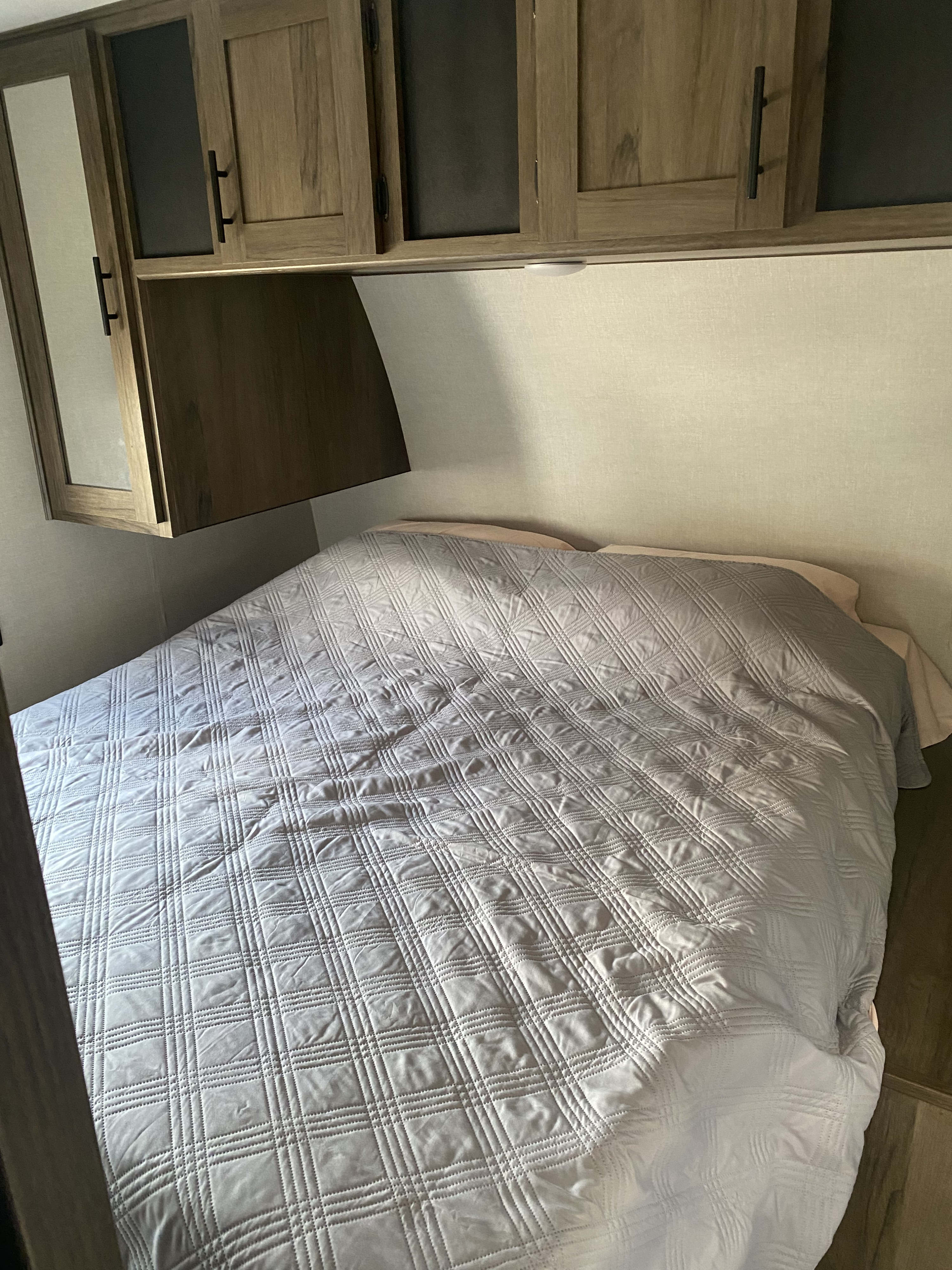Cozy RV rental bedroom featuring a light grey quilted bedspread, overhead wooden cabinets with dark glass inserts, and a side mirror.. Prime Time Avenger 2021