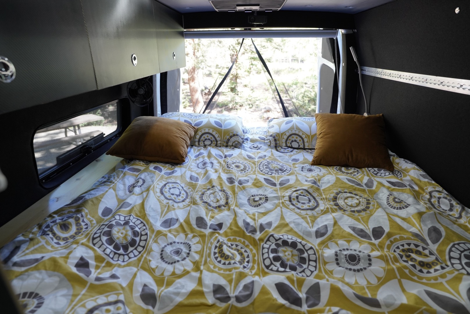 Cozy RV rental bed with yellow, white & grey patterned bedding, brown pillows, and a forest view. Perfect for camper travel.. Mercedes-Benz Sprinter 2021