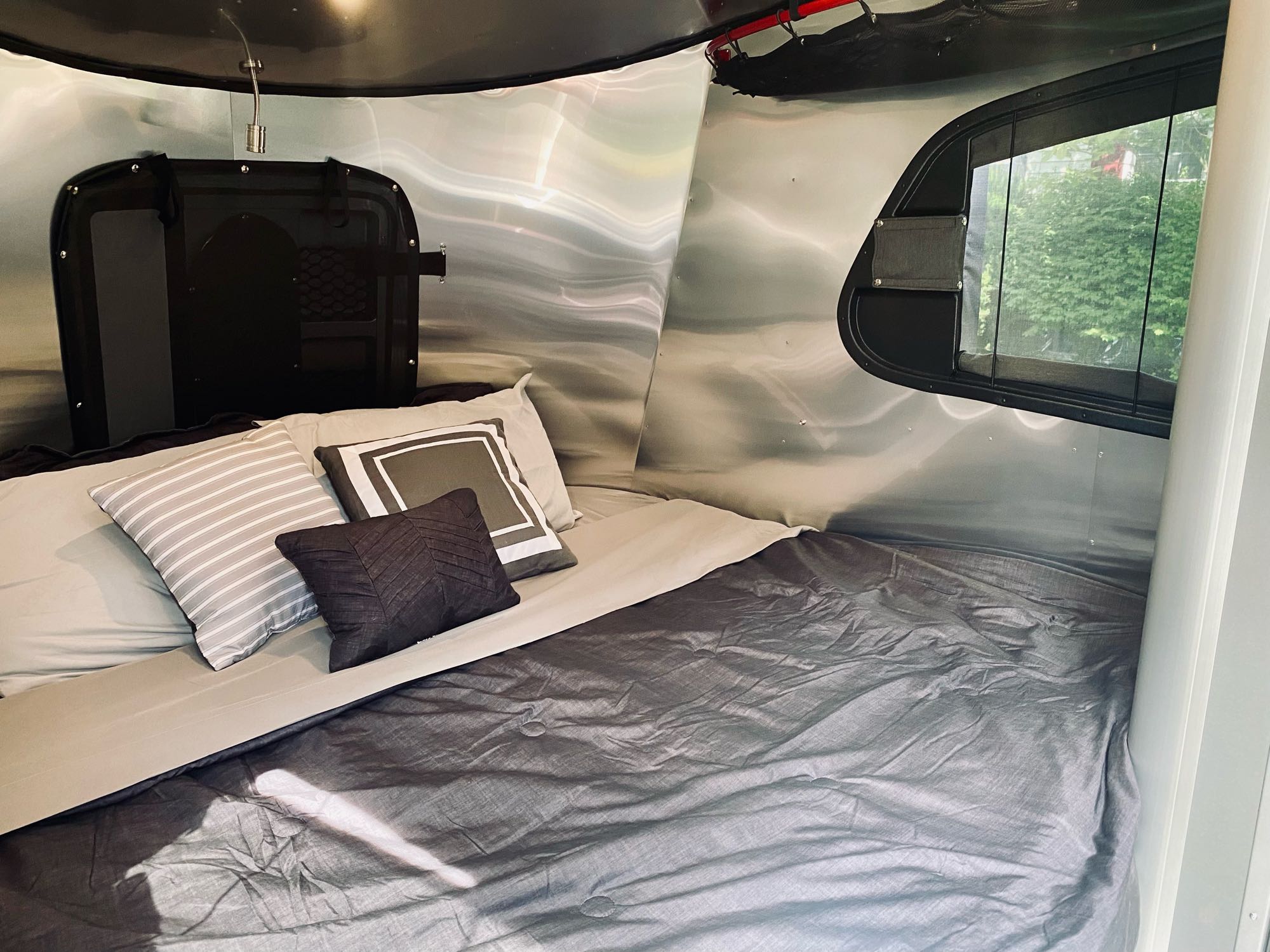 Comfortable sleeping area inside an rv rental, featuring a bed with grey bedding, accent pillows, metallic walls, and a window.. Airstream Basecamp 2021