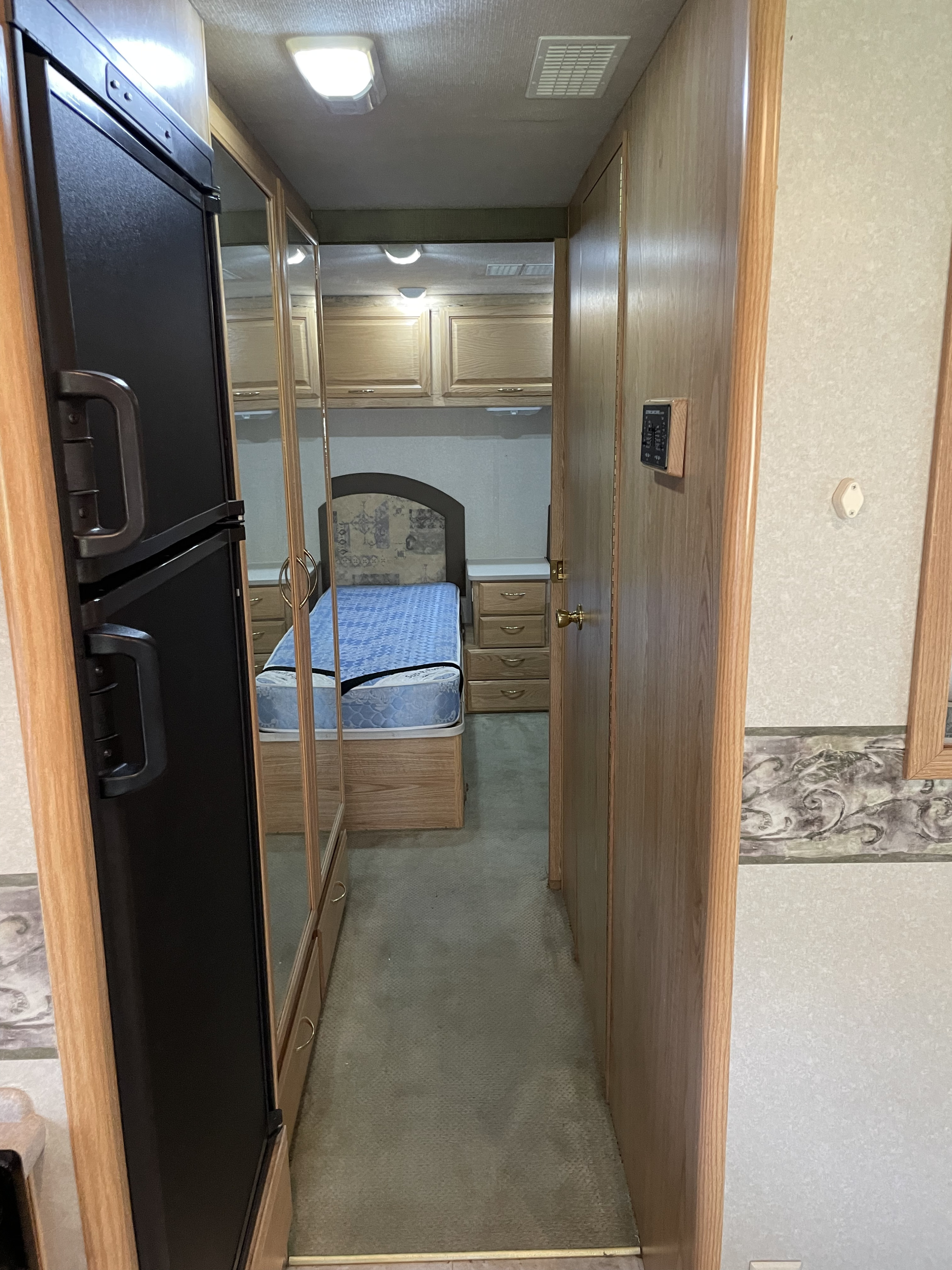 Inside an RV rental: a hallway view with a black refrigerator left, mirrored closet reflecting the bed, and a door right. Carpeted floor, light wood trim.. Bounder Bounder Motorhome 2002