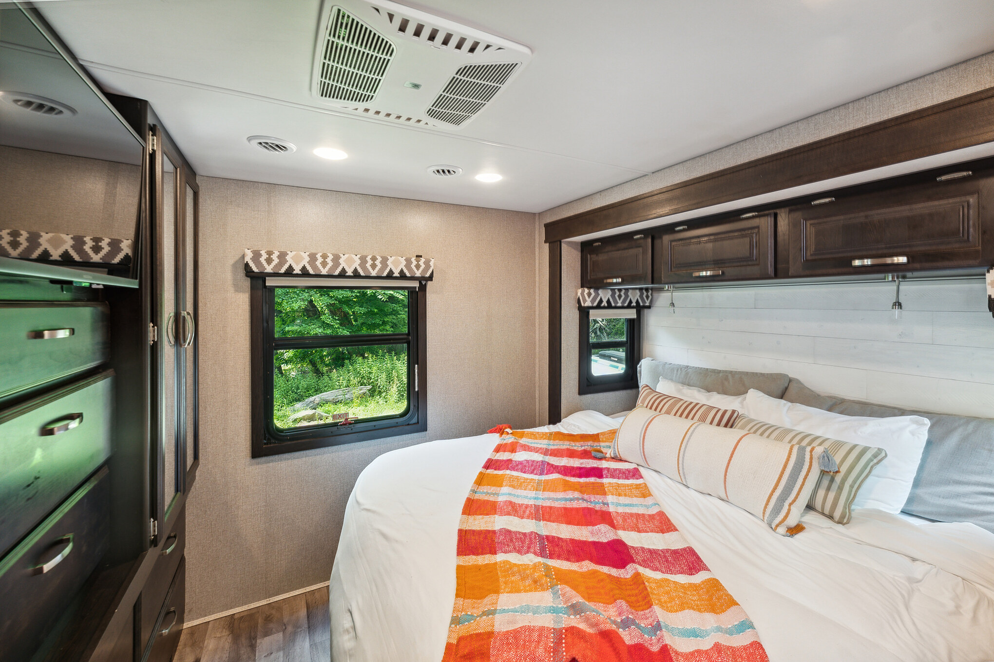 This RV rental bedroom features a comfy bed with a colorful blanket, overhead cabinets, and large windows revealing nature.. Jayco Precept 2021