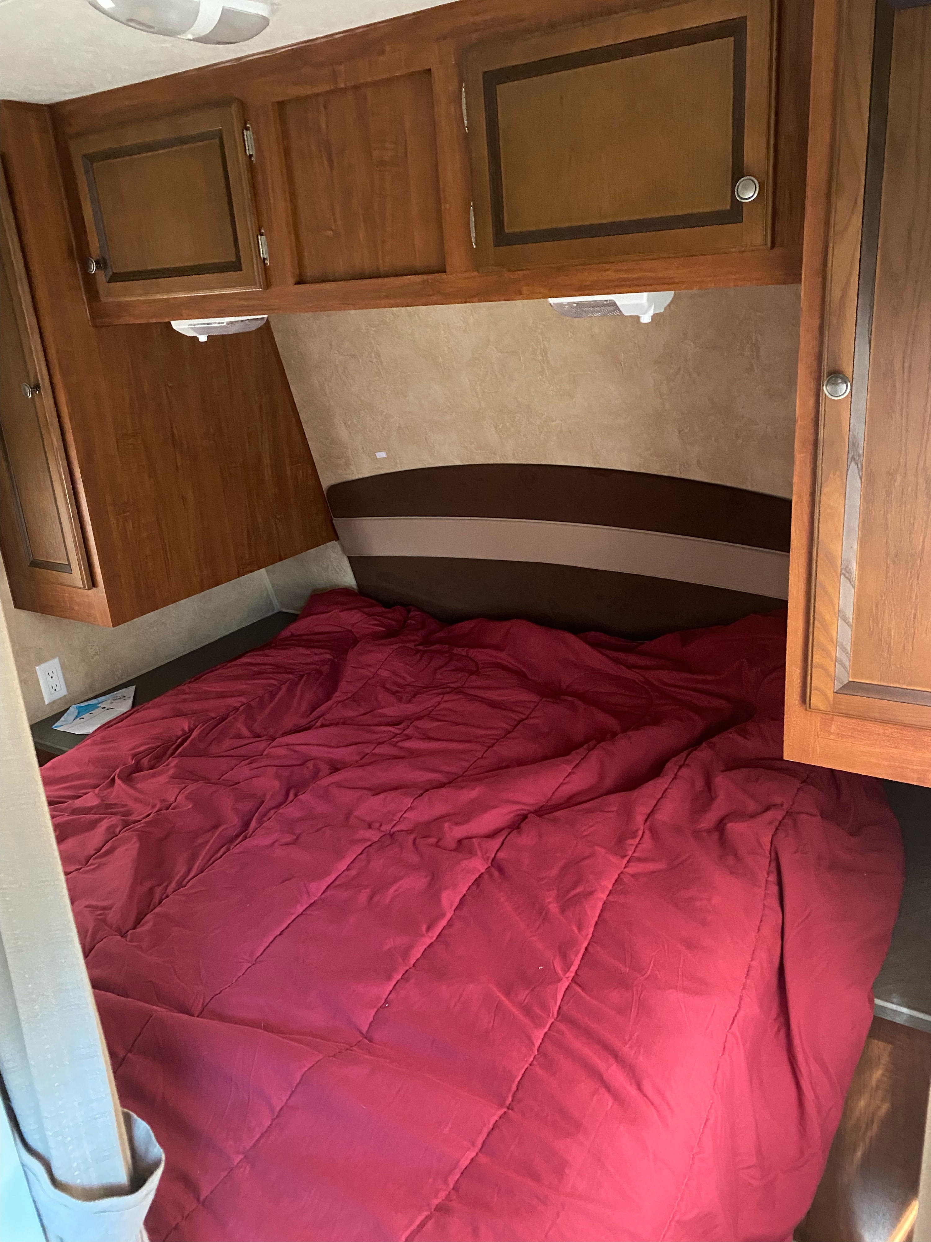 Inside an RV rental, a compact bedroom features a red comforter on the bed, overhead wooden cabinets, and a cushioned headboard.. Jayco Jay Flight 2012