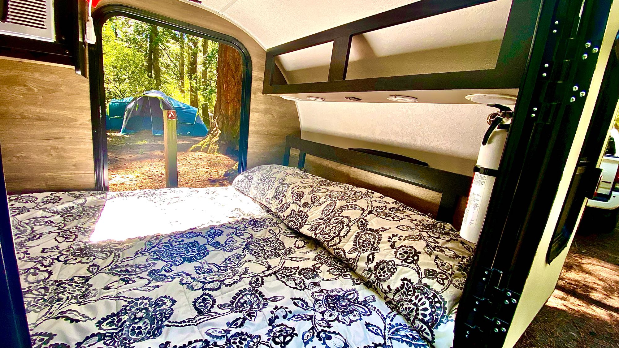 RV rental interior: a comfy bed overlooks a peaceful forest campsite with a blue tent. Ideal for glamping trips.. Other Other 2022