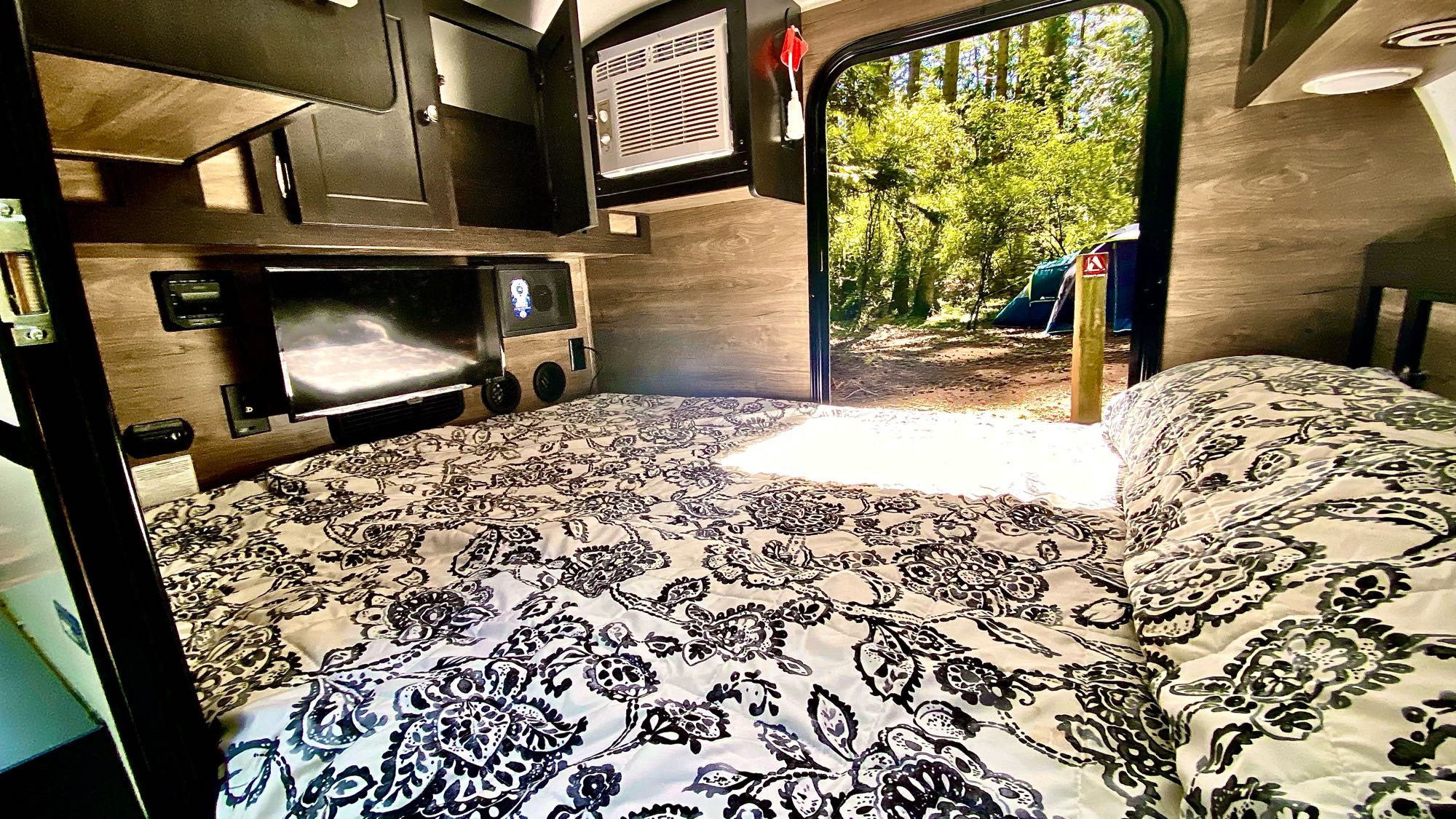 Cozy RV rental interior: bed, TV, & AC. The open door shows a forest campsite with a blue tent & wooden post.. Other Other 2022