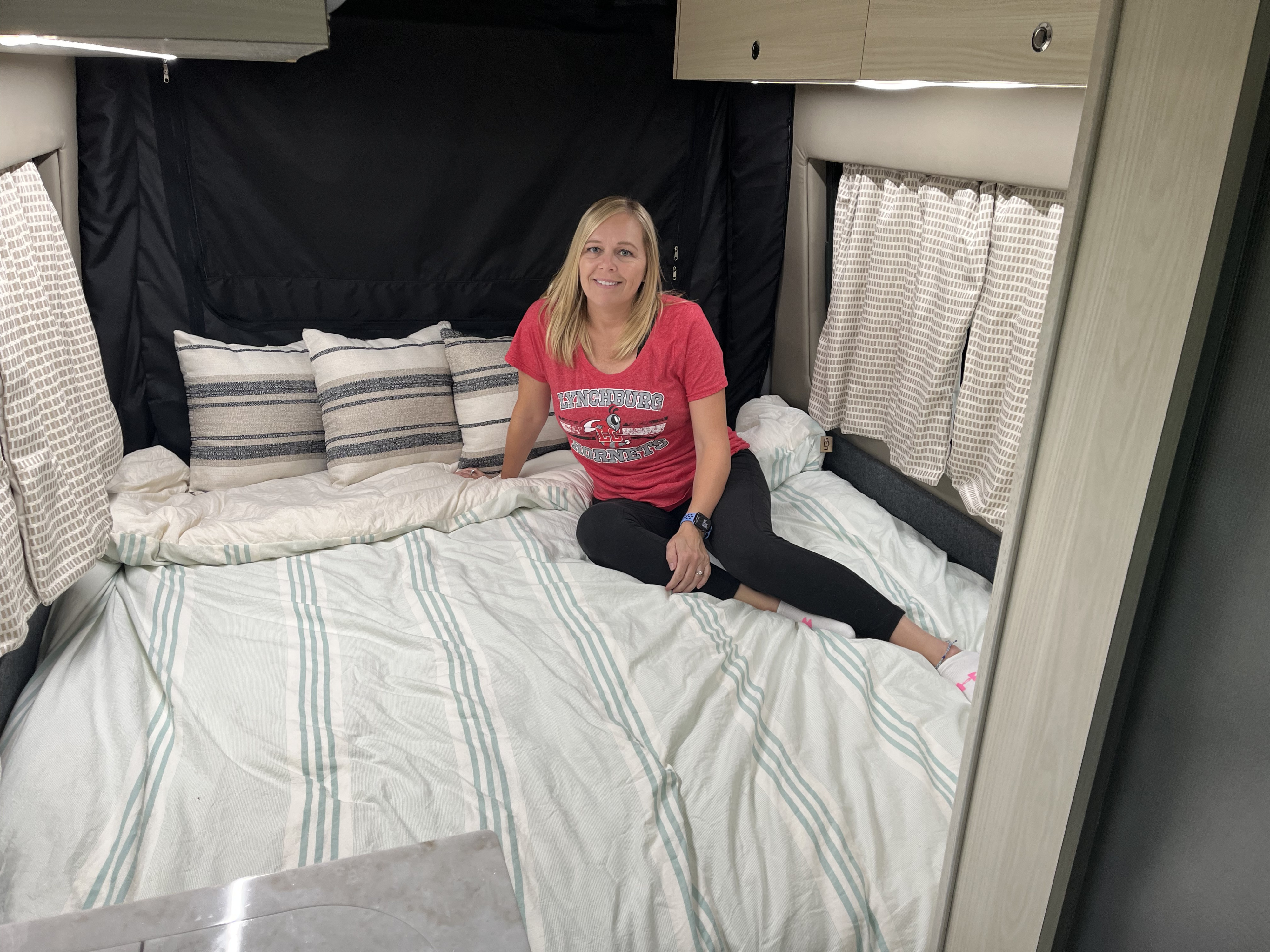 Happy woman sits on a striped bed in a cozy RV rental, showing the comfortable interior.. Tiffin Motorhomes Cahaba 2021