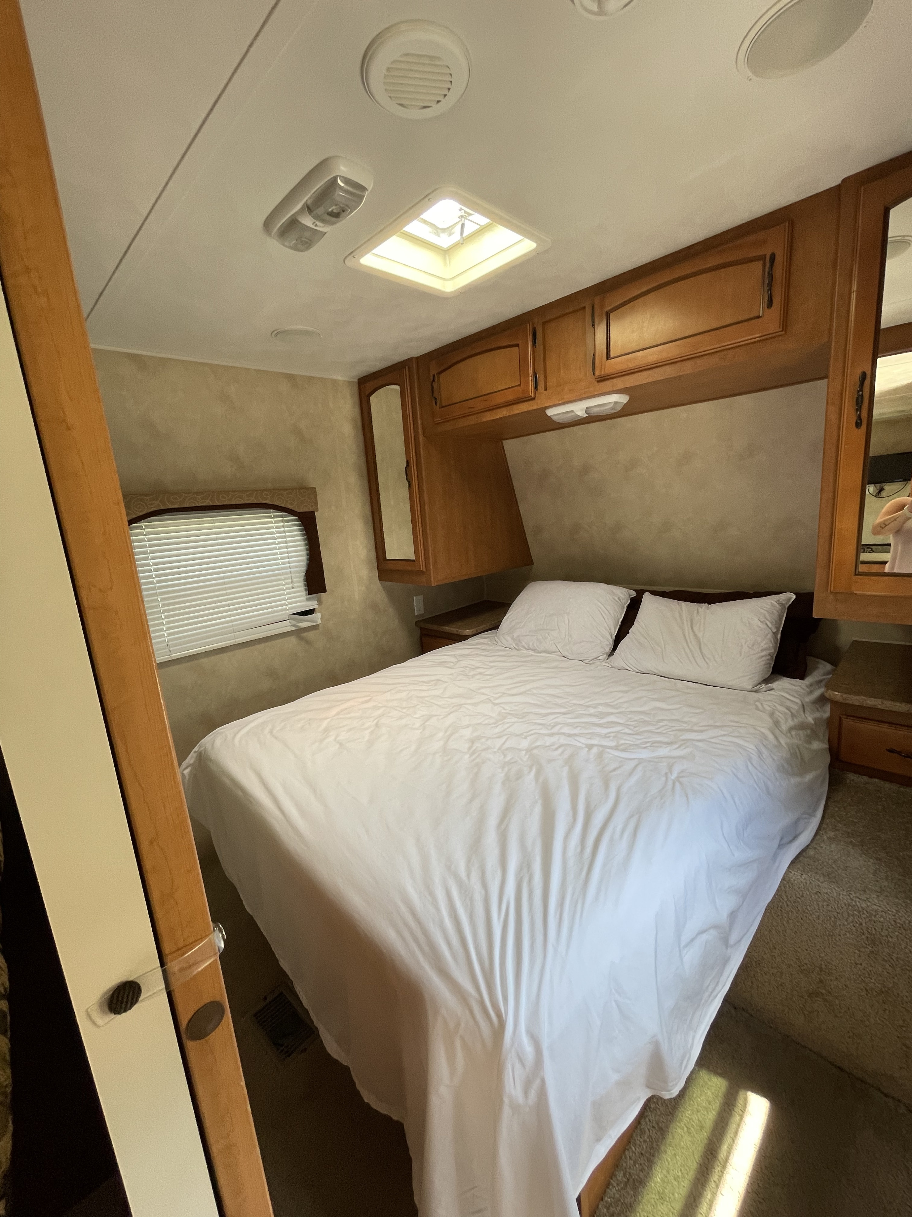 Cozy RV rental bedroom featuring a bed with white linens, overhead wooden cabinets, a window with blinds, and a ceiling skylight.. Forest River Palomino Puma 2012