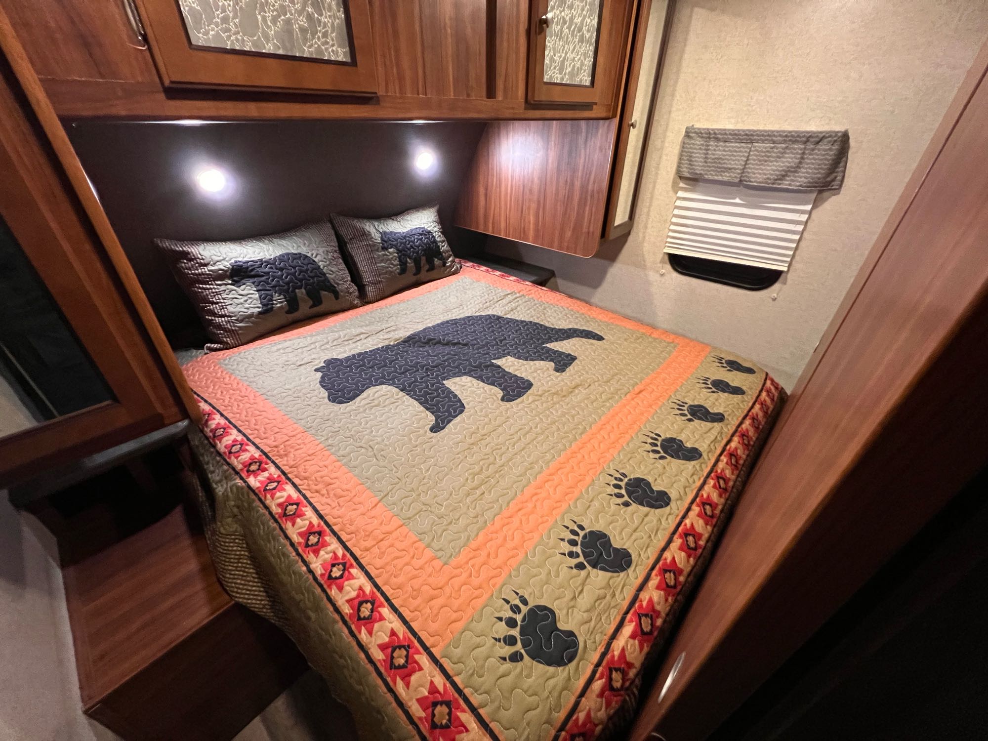Cozy RV rental bedroom with a bear-themed quilt and pillows, overhead storage, reading lights, and a window blind.. Heartland RVs Mallard 2018