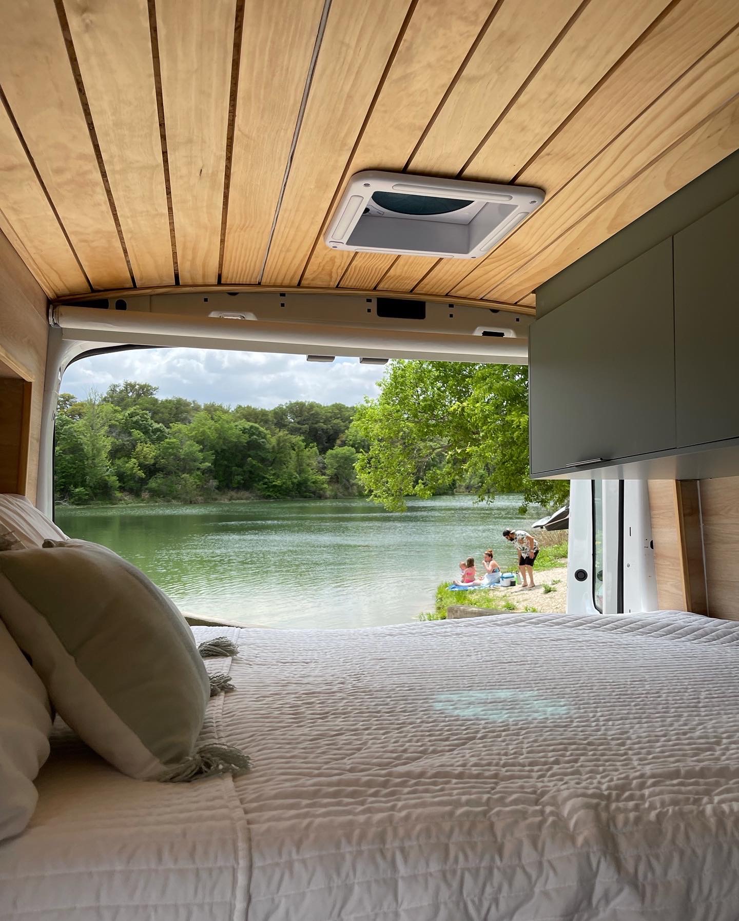Relax by the lake! This cozy rv rental offers a stunning view from bed, with people enjoying the water's edge and lush trees.. Ford Transit 2019