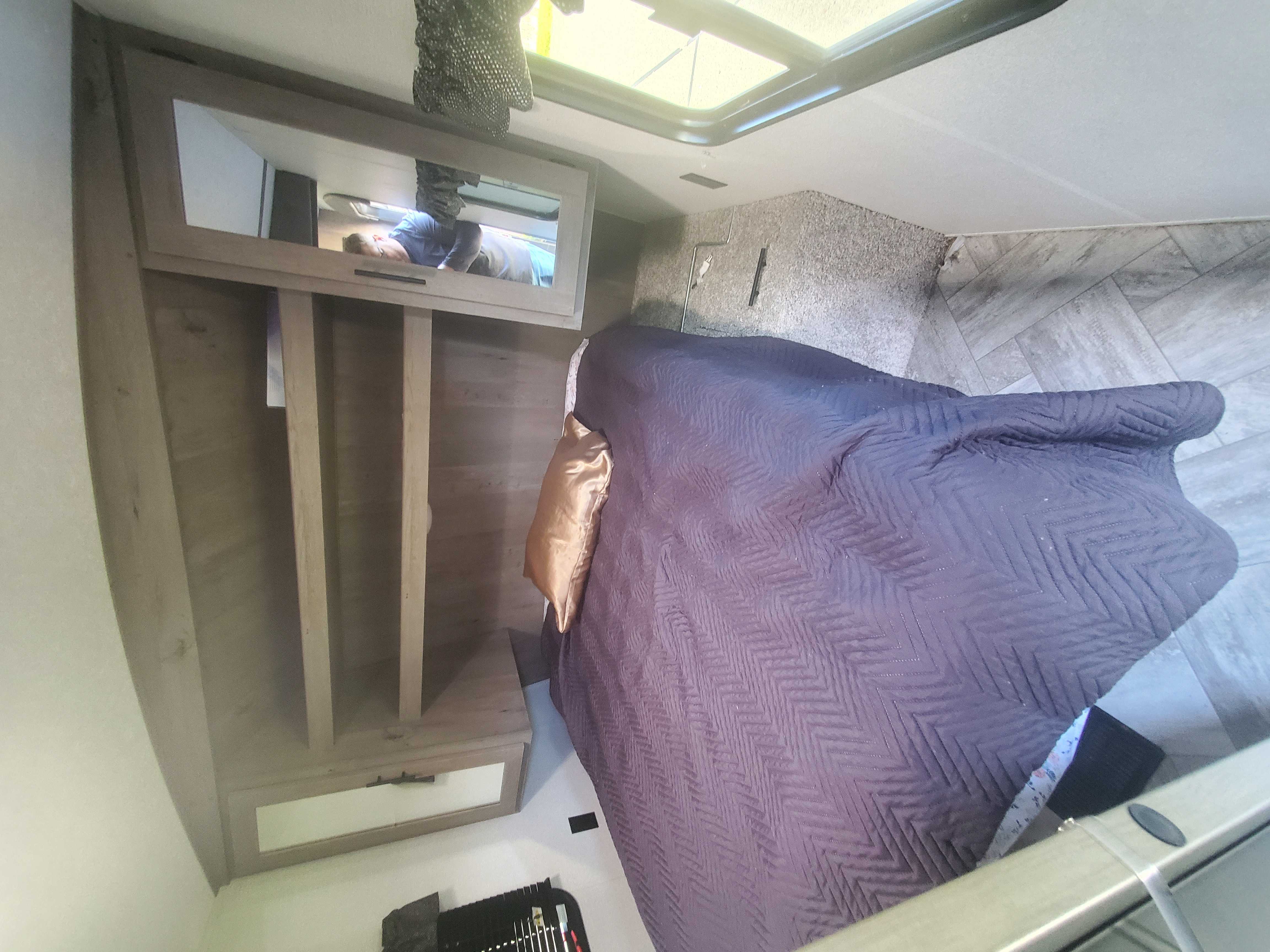 RV rental bedroom interior featuring a bed with purple blanket, mirrored cabinet, and overhead skylight. Person visible in reflection.. Keystone Springdale 2021
