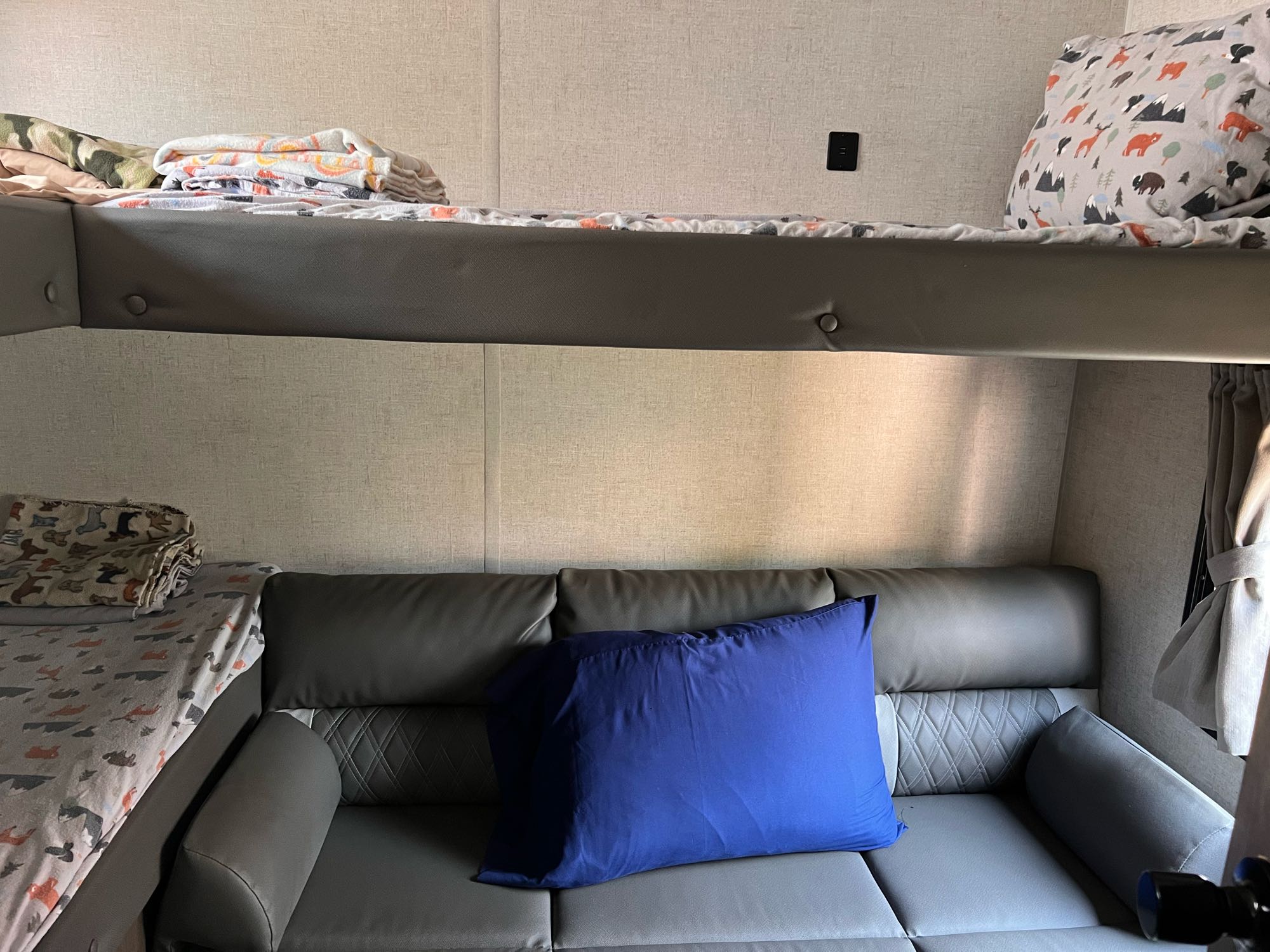 Inside an RV rental: a cozy upper bunk bed with patterned sheets and a versatile lower area, functioning as a bunk or sofa.. Forest River Wildwood X-Lite 2021