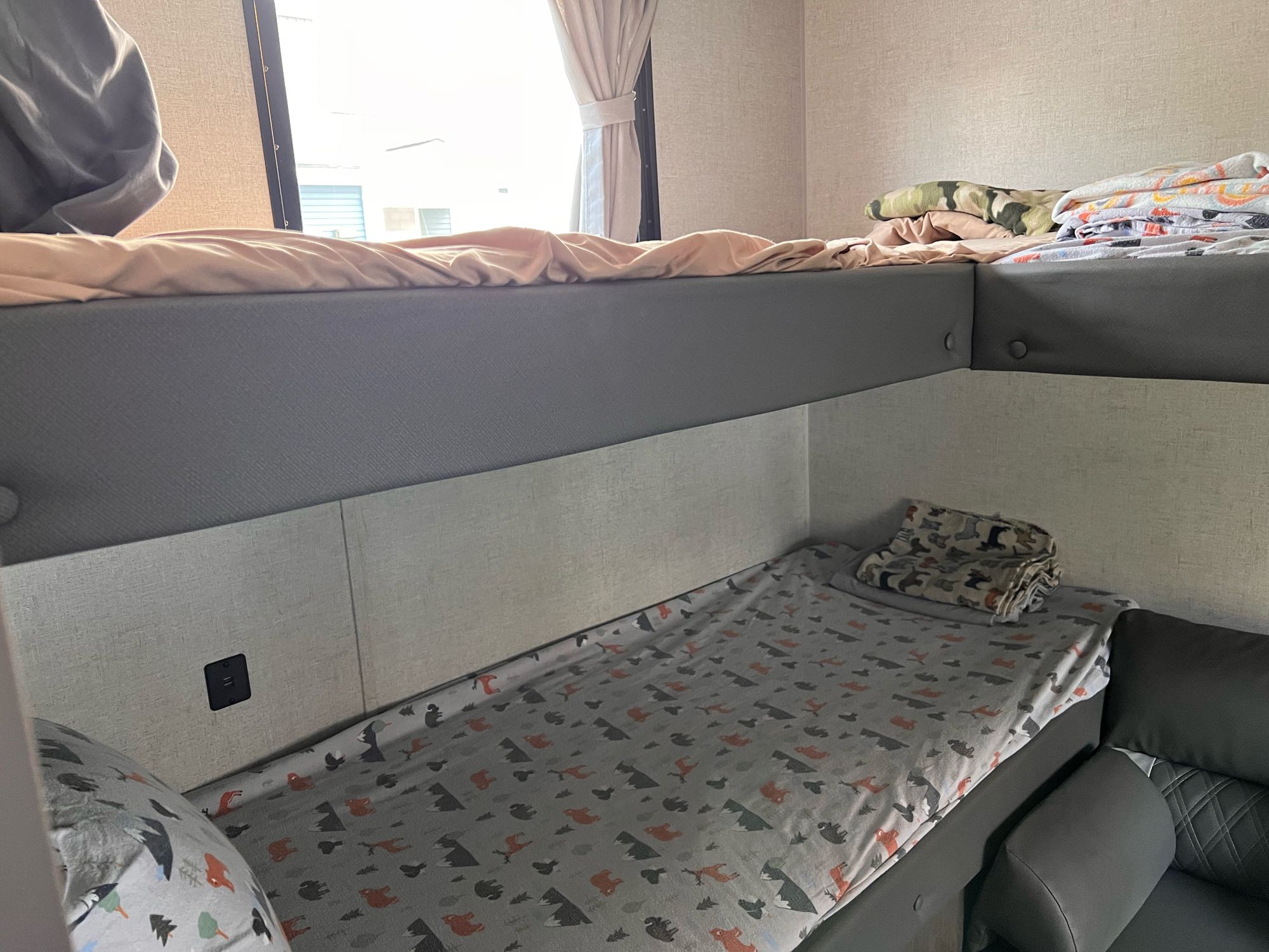 Inside an RV rental: cozy bunk beds with patterned and solid bedding, a window, and a wall outlet offer compact sleeping.. Forest River Wildwood X-Lite 2021
