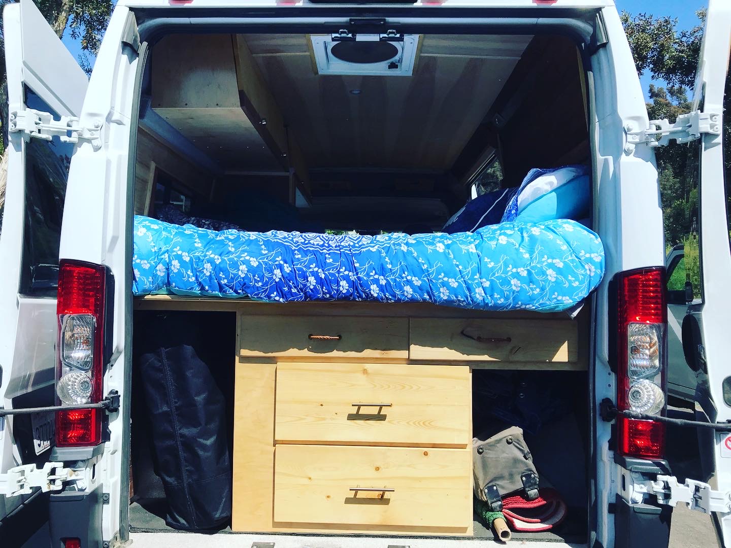 Back of an RV rental van, revealing a cozy blue-patterned bed above custom wooden storage. Ideal for road trips & camping.. RAM Promaster 2018