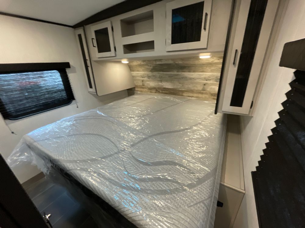 Spacious, bright RV rental bedroom with a new, plastic-covered mattress, modern overhead cabinets, and window.. Heartland RVs Mallard 2022