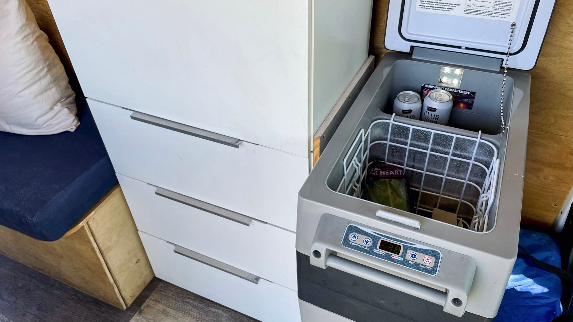 RV rental interior: A portable fridge stocked with drinks and food sits next to white storage drawers and a built-in bed.. Dodge Sprinter 2017