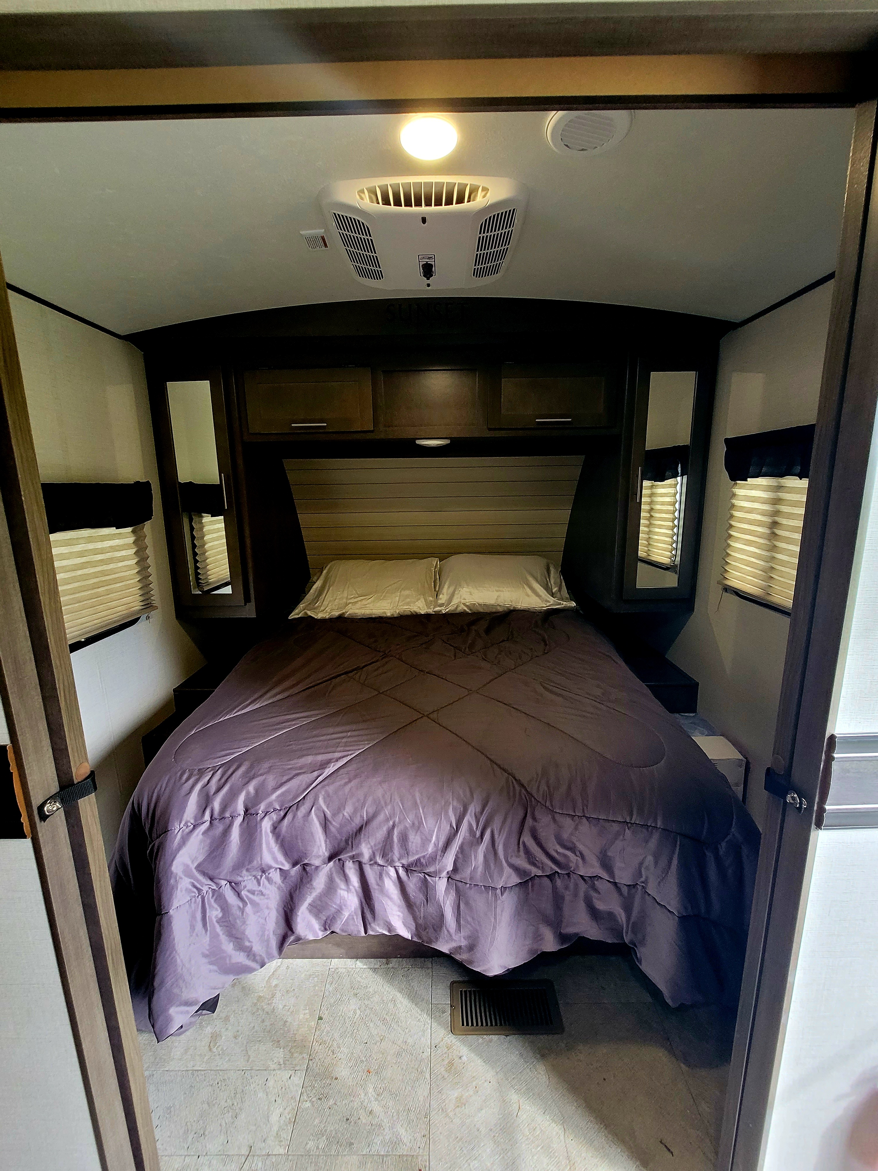 This RV rental bedroom interior features a cozy bed with purple bedding, overhead storage, an AC unit, and windows.. Crossroads RV Sunset Trail Super Lite 2018