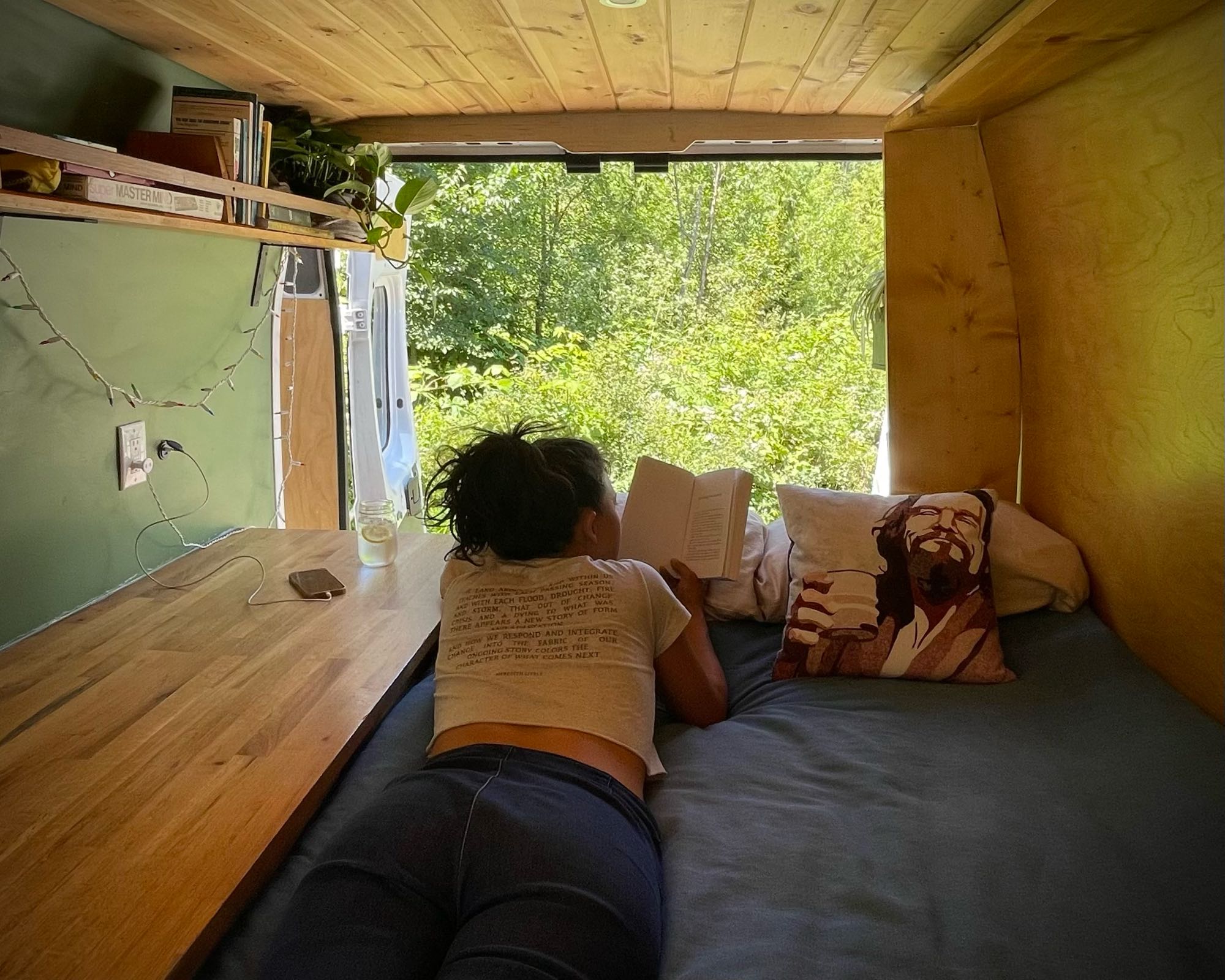 Cozy RV rental: A person reads a book inside a camper van, doors open to a serene forest view. Perfect van life escape.. Dodge Sprinter 2017
