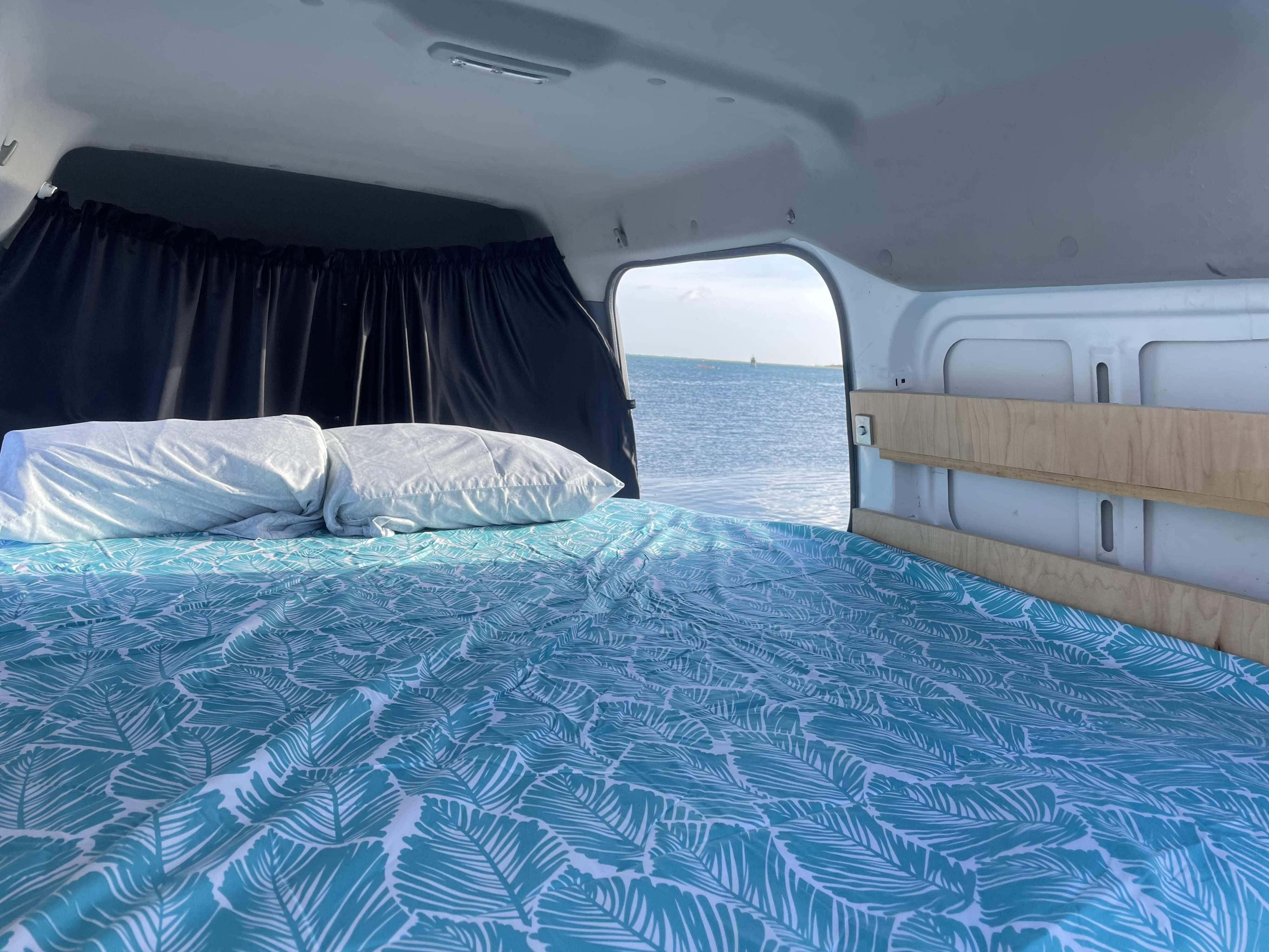 Inside a bright RV rental: a bed with a teal leaf-patterned duvet and white pillows overlooks a calm ocean. Ideal campervan adventure.. Ford Transit Connect 2010