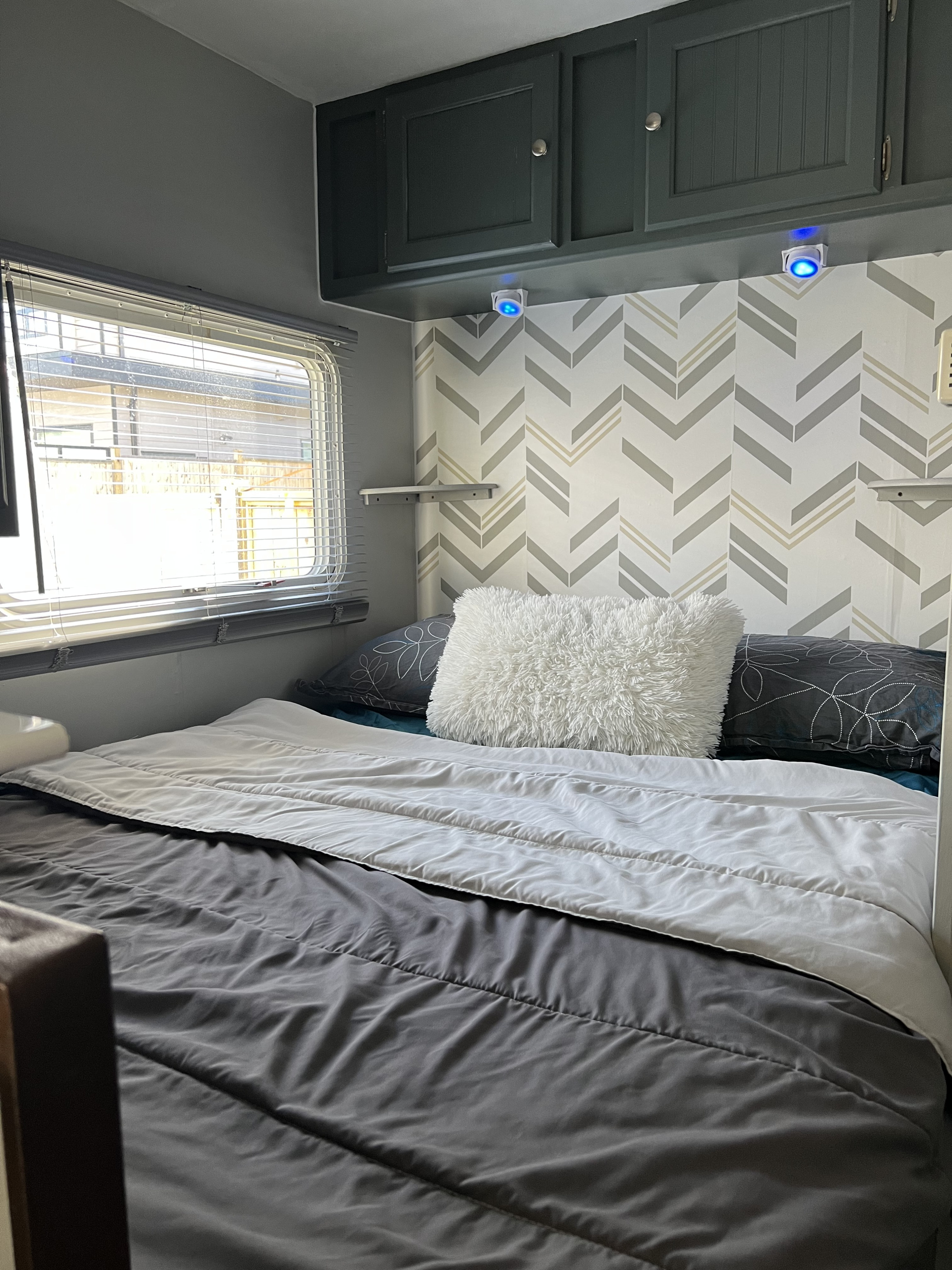 An RV rental bedroom showcasing a bed with gray bedding, dark cabinets, blue lights, and a gray/beige chevron wall.. Dutchmen Sport 2004