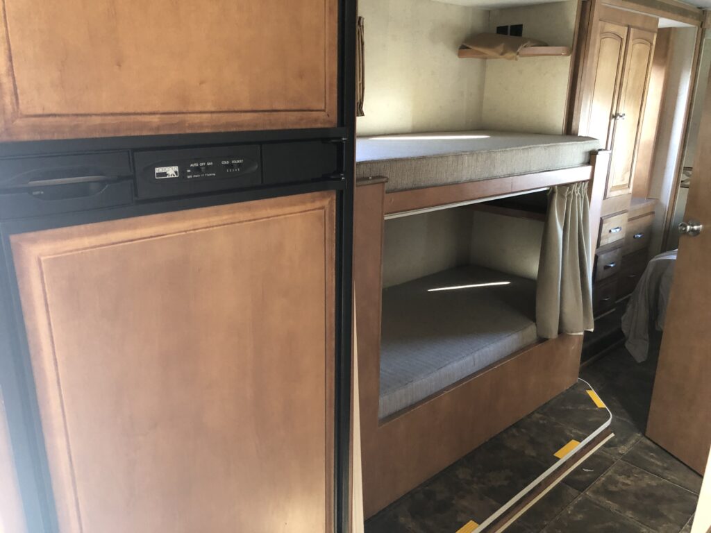 RV rental interior showing a wood-paneled refrigerator, bunk beds with mattresses, and wooden cabinetry. A compact travel space.. Winnebago Chalet 2014