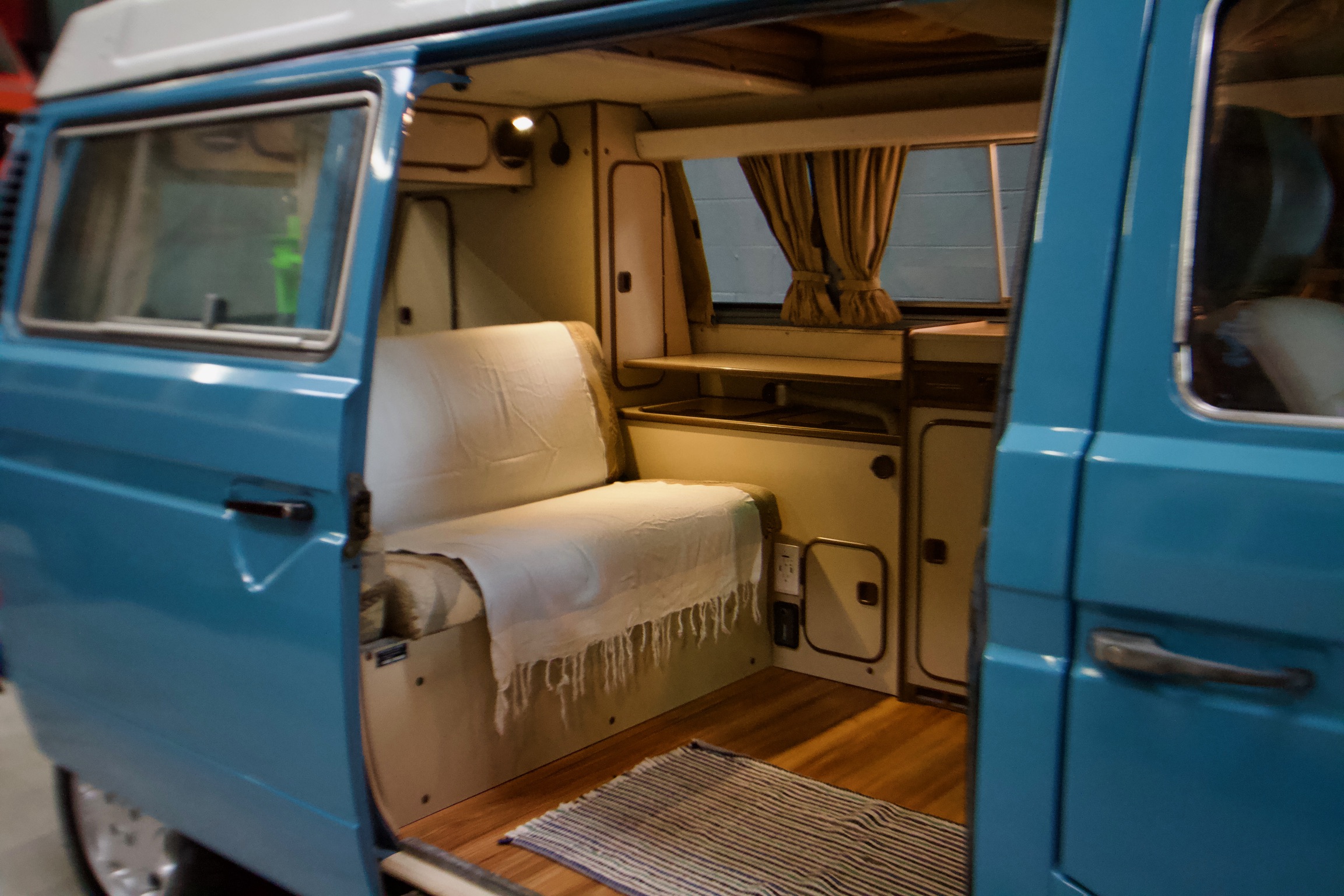 Open side door reveals a cozy blue camper van interior, ideal for an RV rental. Features a bench seat, wood floor & curtains.. Volkswagen Westfalia 1982