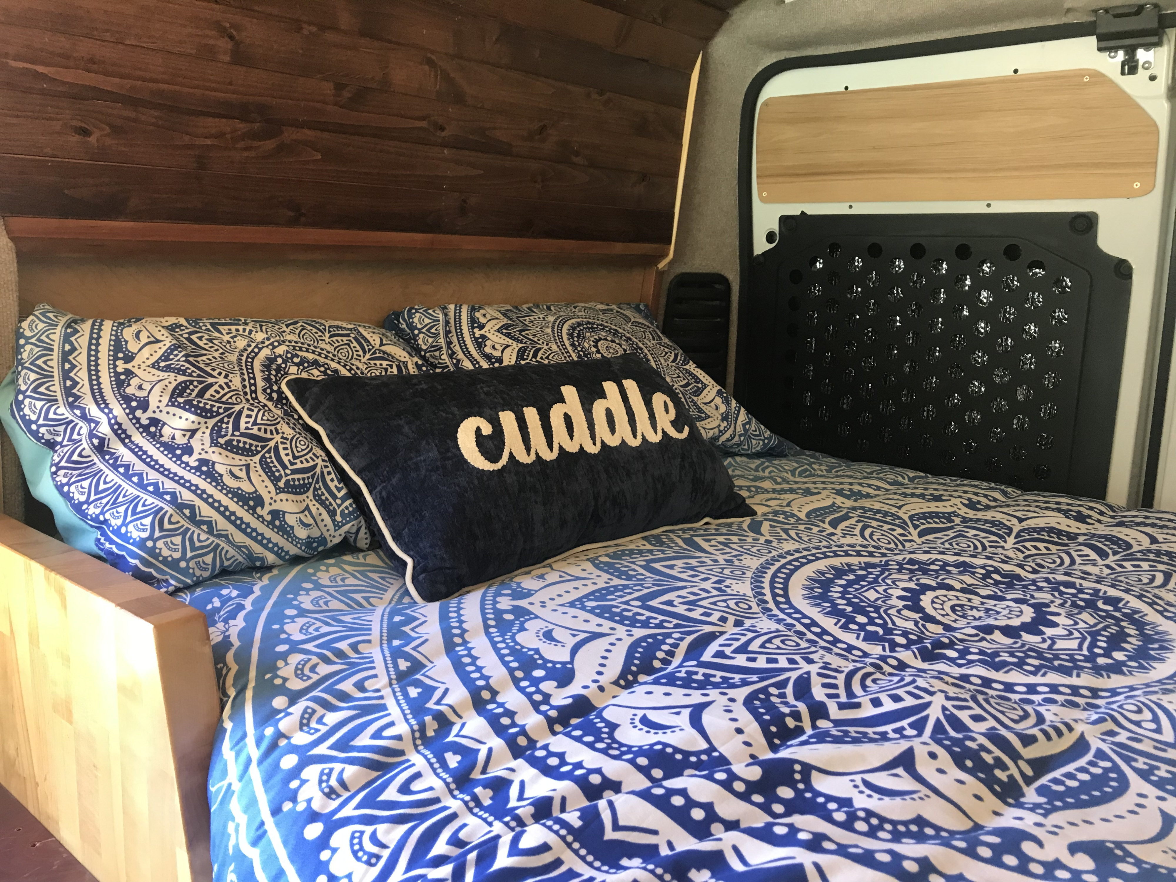 Cozy RV rental bed with blue mandala bedding and "cuddle" pillow. Features warm wooden interior and details.. RAM Promaster 2018
