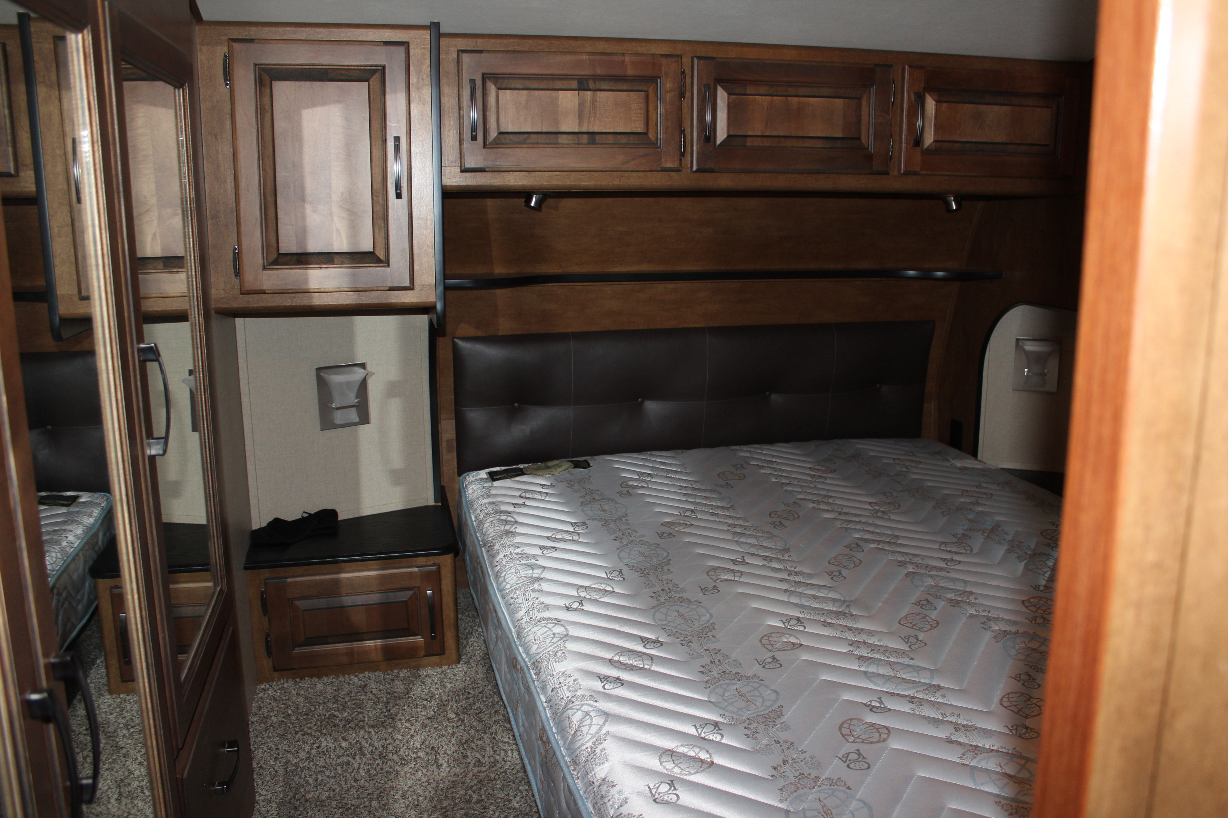 RV rental bedroom featuring a bed, padded headboard, and overhead wood cabinets. Compact and functional.. Grand Design Reflection 2015