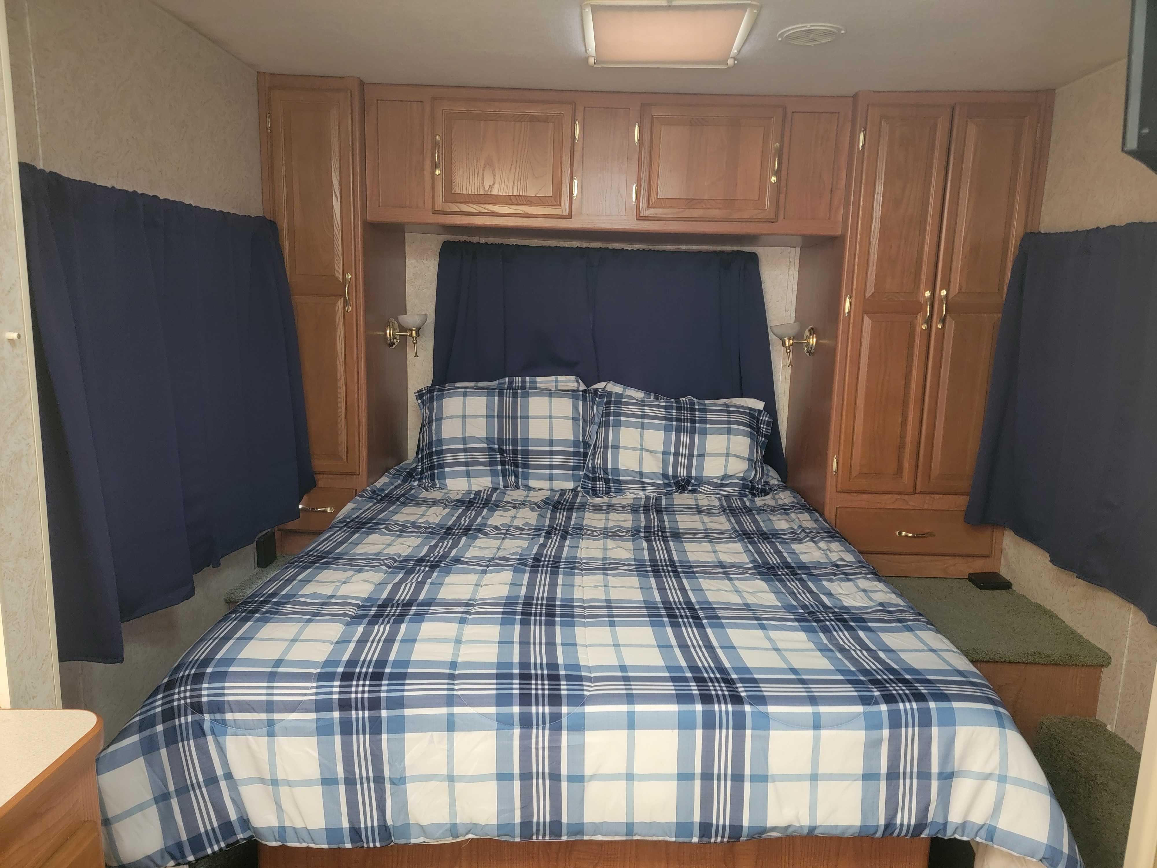 Cozy RV rental bedroom with a blue plaid bed, overhead wood cabinets, side curtains, and accent lighting.. Coachmen Santara 2003