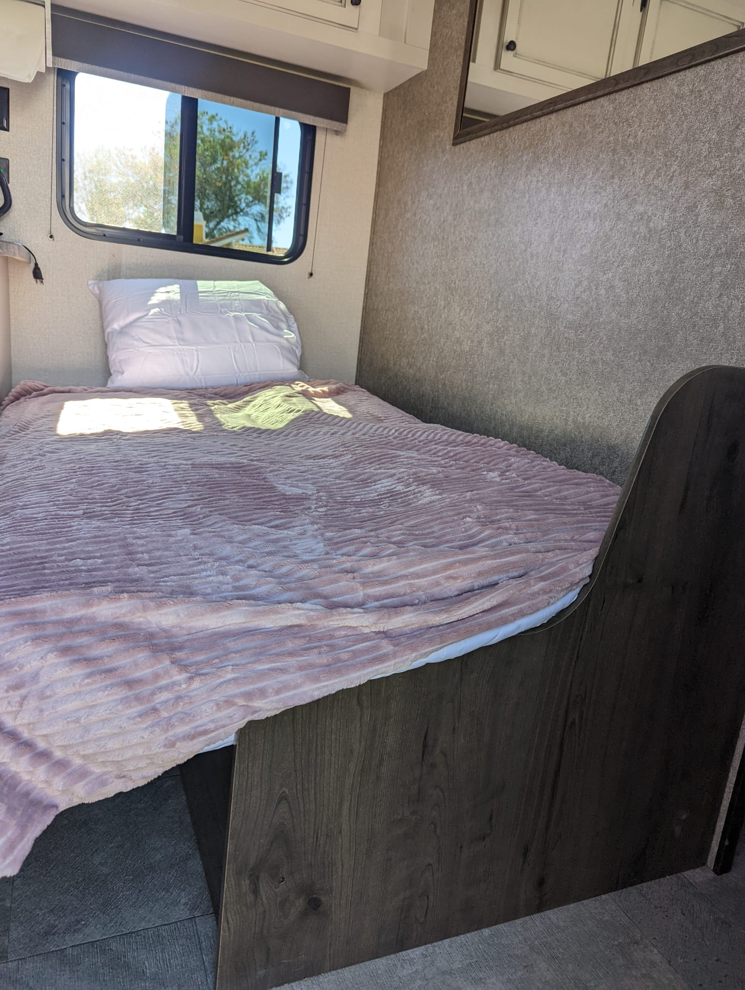Bright RV rental bedroom features a comfy single bed with a pink blanket, a window view, and textured walls.. Highland Ridge RV Open Range 2021