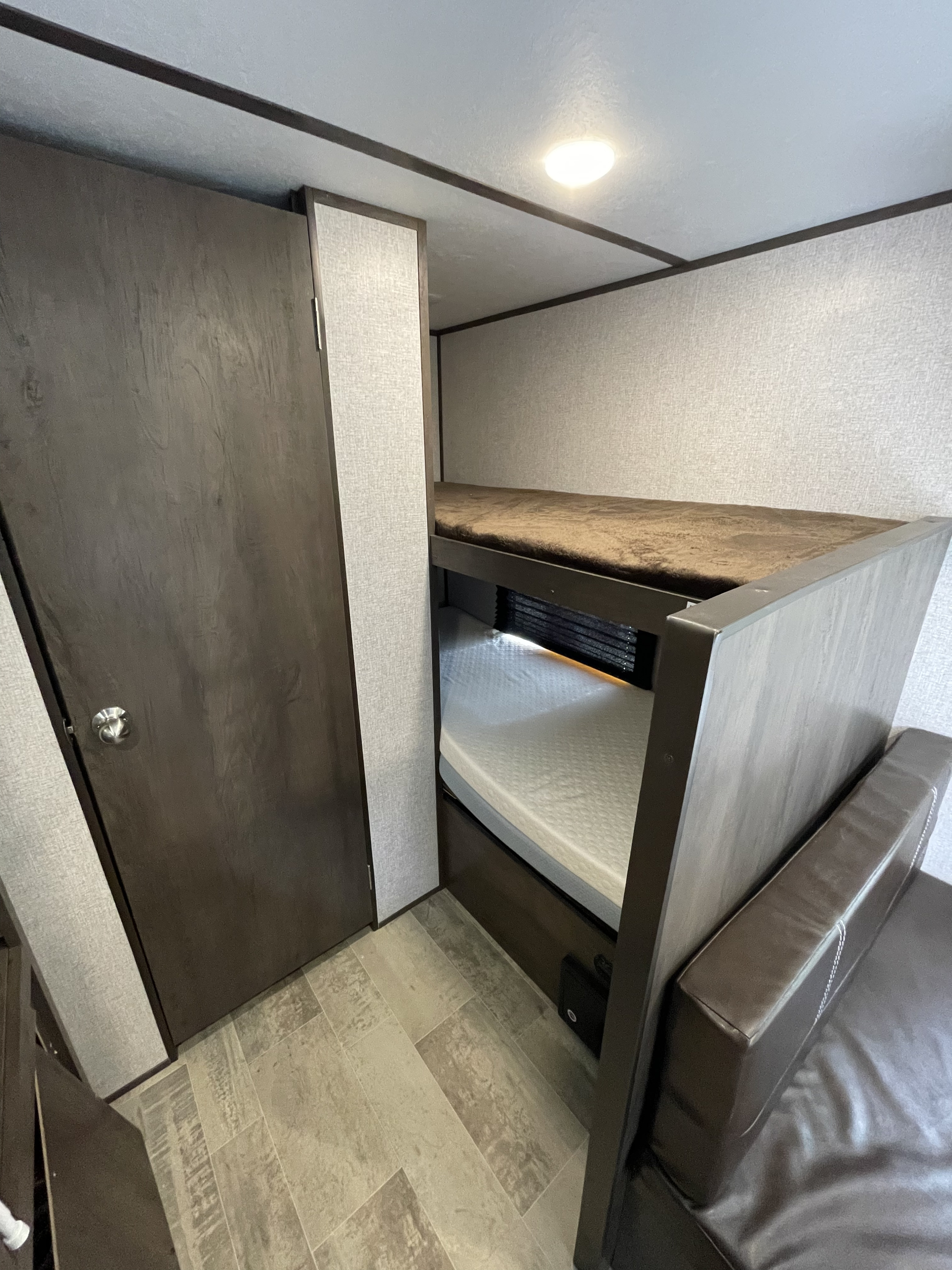 Inside an RV rental: dark wood bunk beds, a closed door, brown leather sofa. Light textured walls, tiled floor.. Dutchmen Coleman Lantern LT 2021
