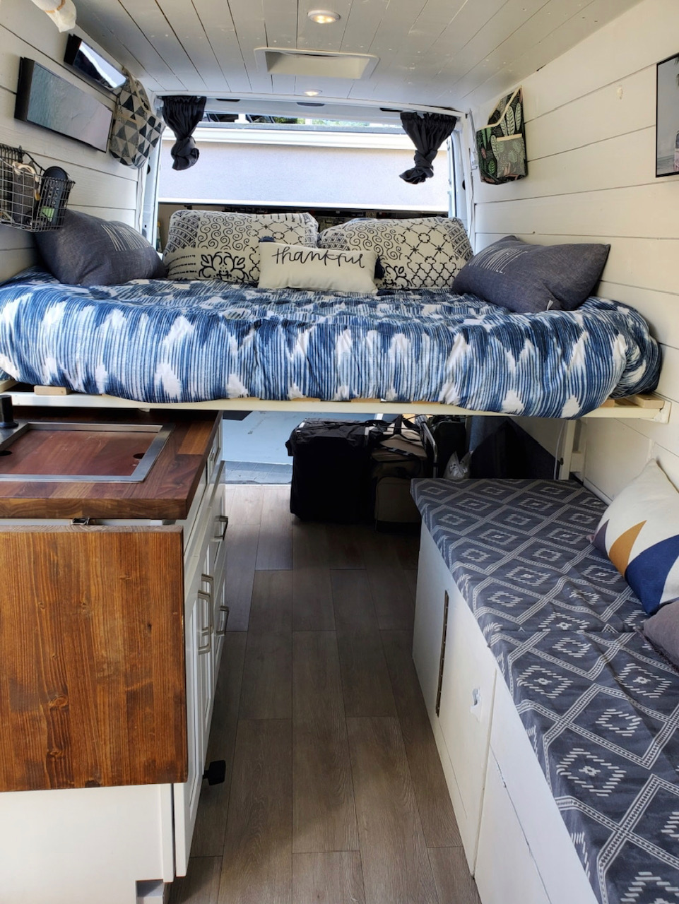 Cozy RV rental interior featuring an elevated bed with blue bedding, a compact kitchen, and side bench seating.. Mercedes-Benz Sprinter RV Motorhome Campervan 2016