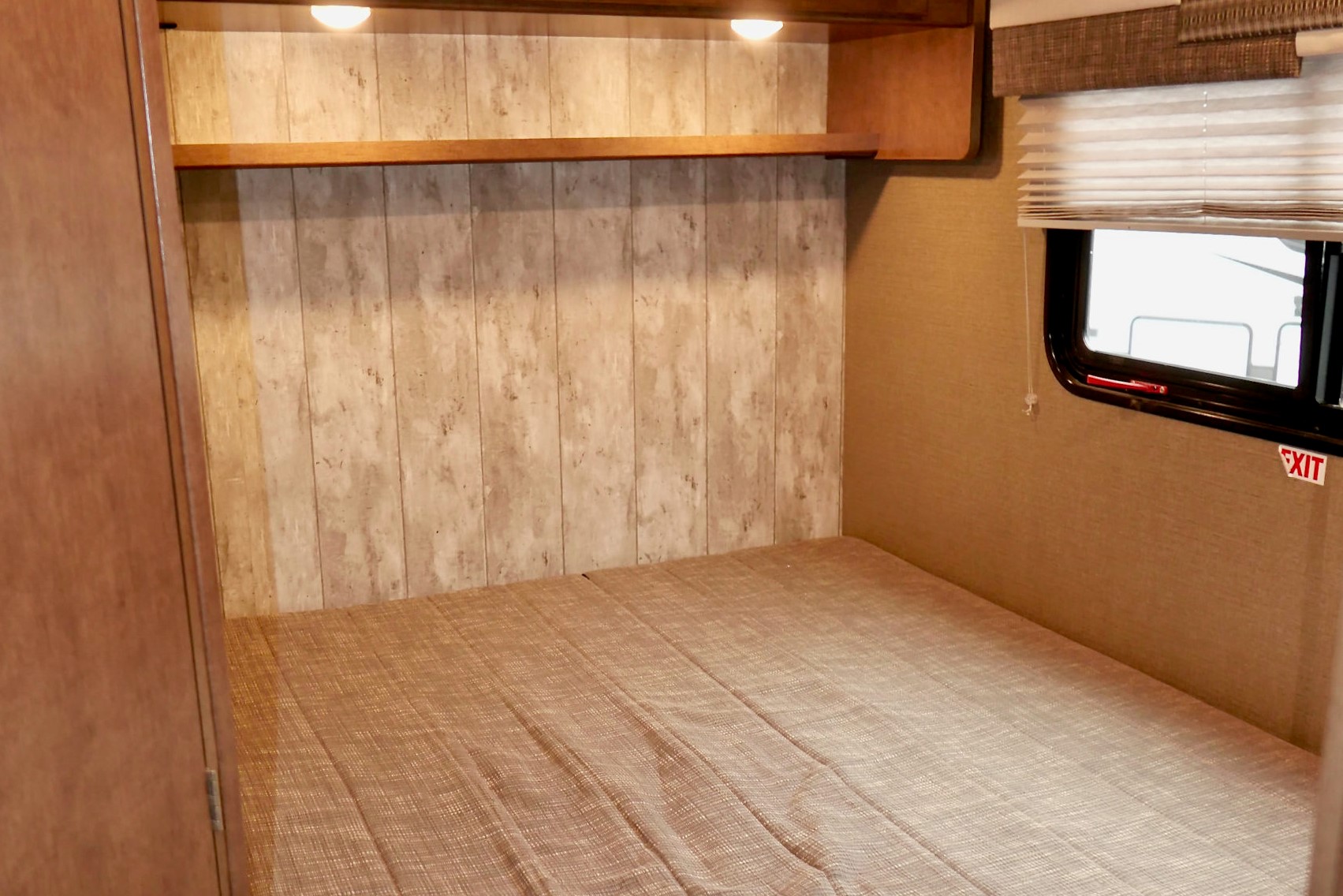 RV rental bedroom: a bed with patterned cover, wood-look wall, upper shelf with lights, and a window with an EXIT sign.. Winnebago Outlook 2019