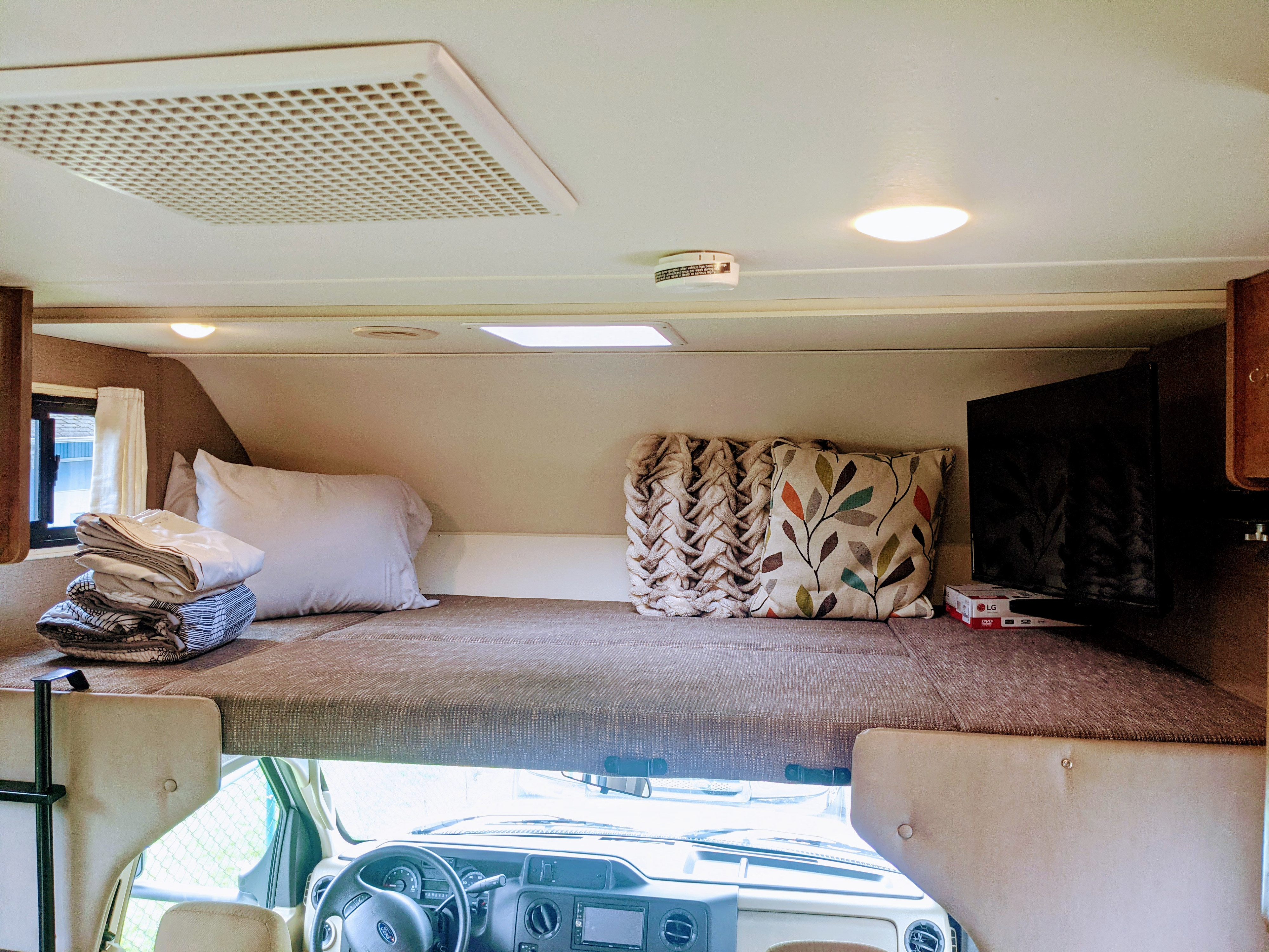 Cozy overhead bunk bed in an RV rental, with bedding, TV, and cab view below. A comfortable sleeping area.. Winnebago Outlook 2019