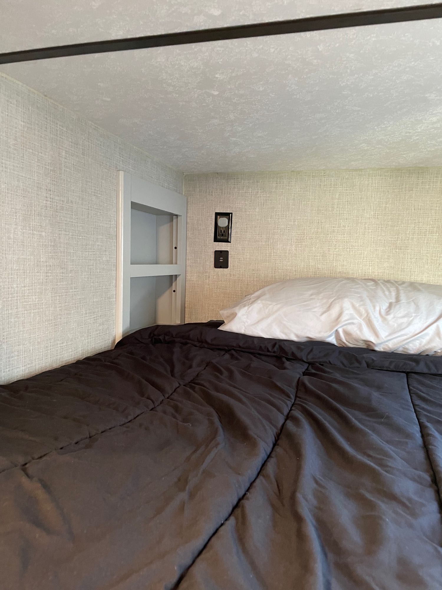 Cozy RV rental bed: bunk with textured walls, built-in shelving, outlets, and a dark comforter.. Keystone Bullet 2020