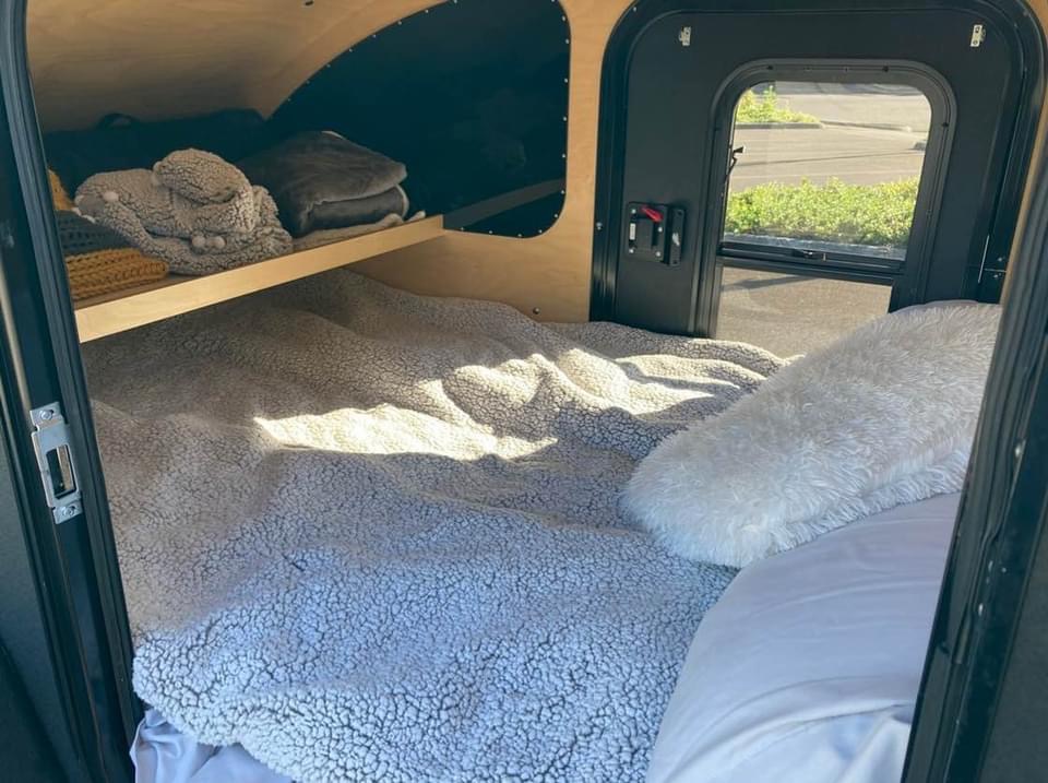 Inside a cozy rv rental: a comfy bed with a sherpa blanket, shelf, and window. Perfect for your next road trip adventure.. Oregon Trail’r DoDrop Alpha 2018