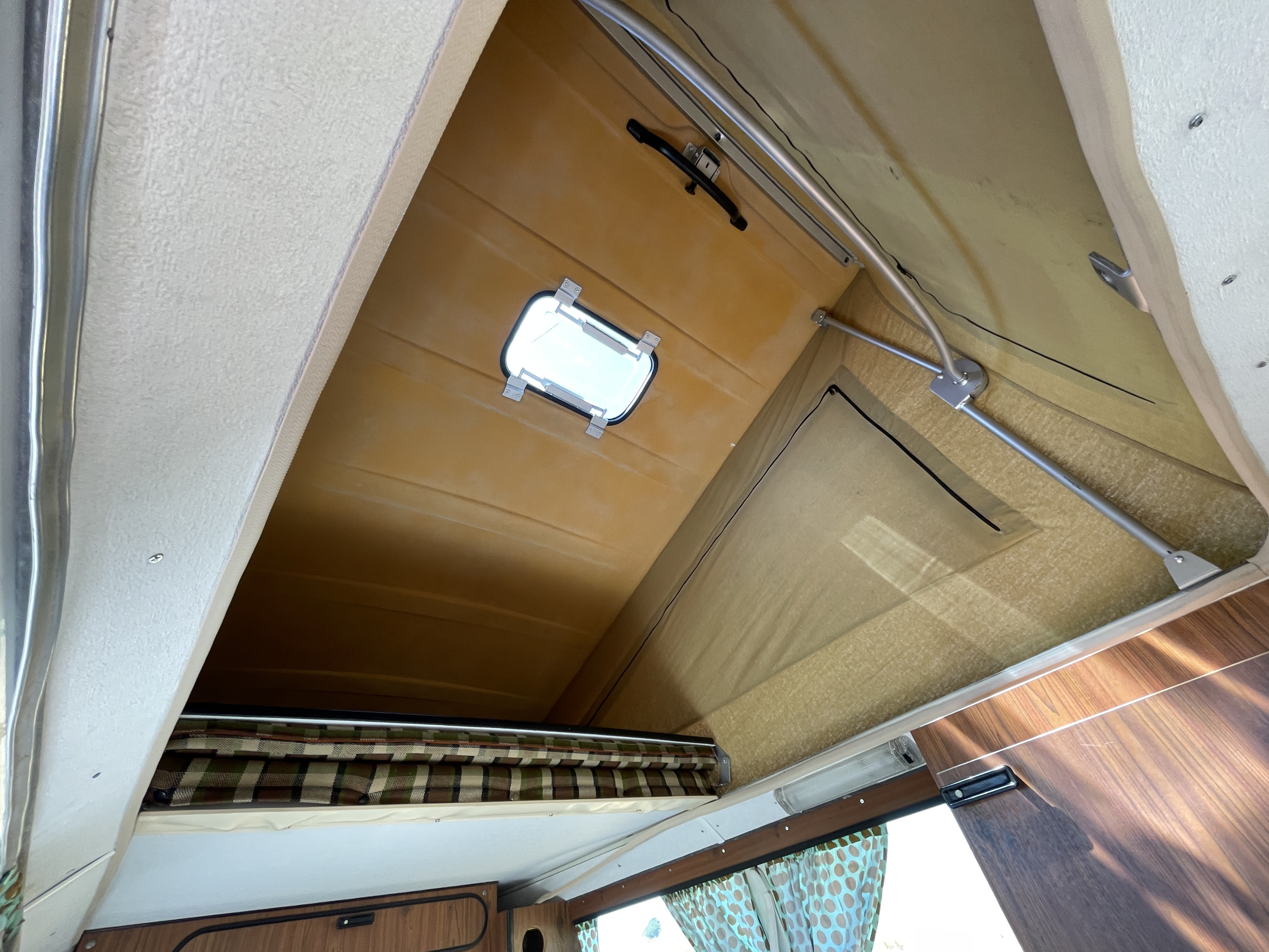 View inside an RV rental with its pop-top roof raised, revealing a window, metal supports, and an upper bunk bed.. Volkswagen westfalia 1976